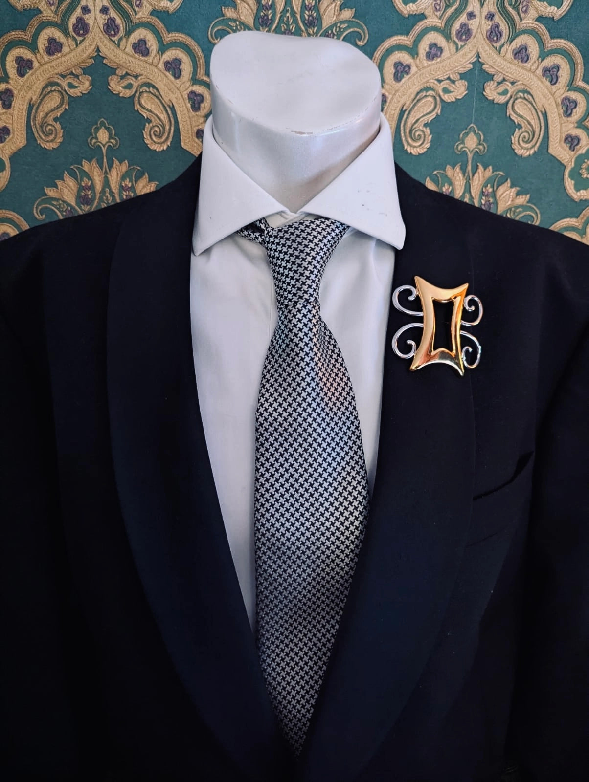 Broochella men's brooches with meaning strong and humble gold metal and silver brooch. Pinned to a men's mannequin wearing a black suit and houndstooth tie. The background is green and gold damask wallpaper.