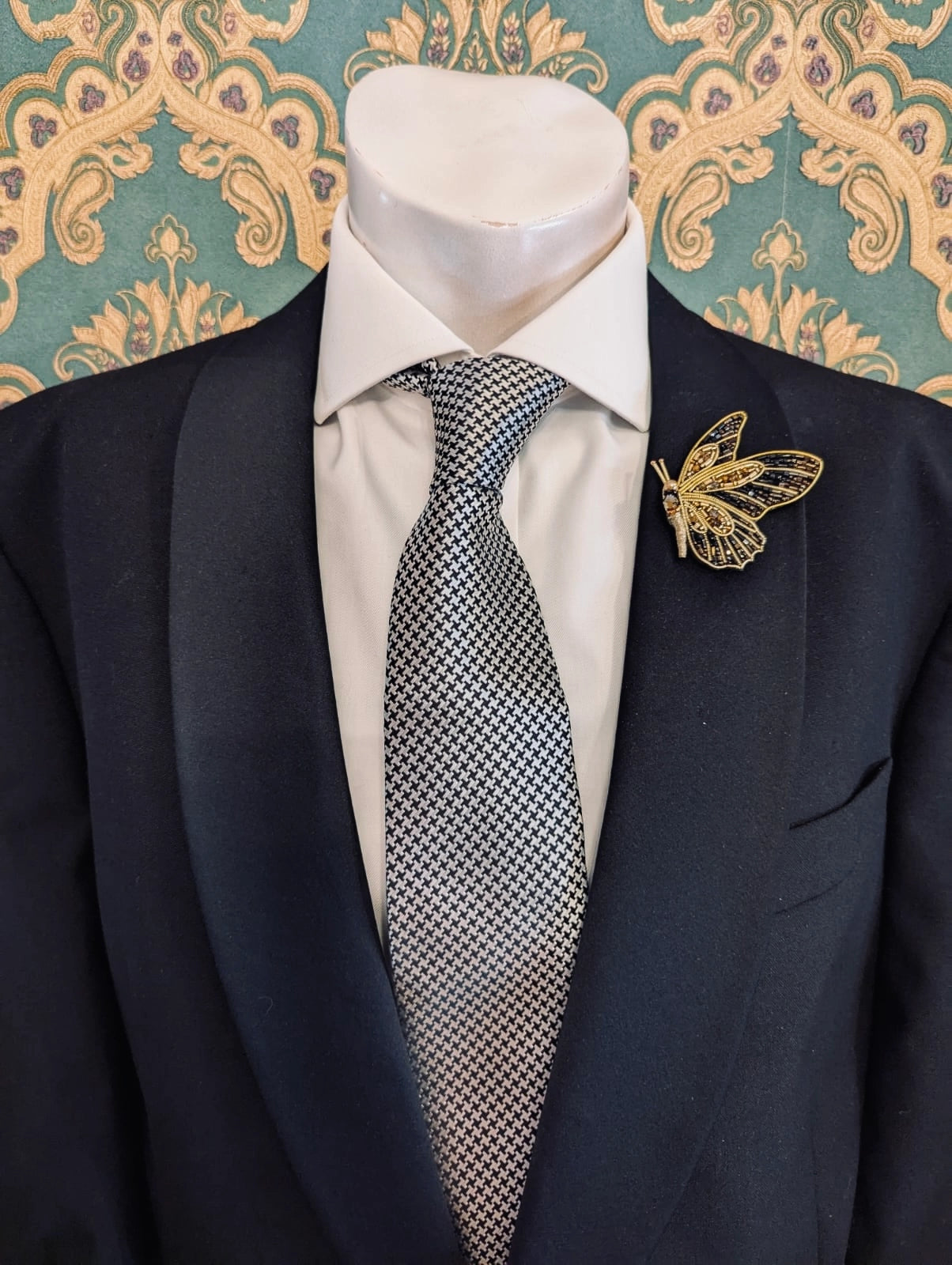 Broochella men's hand beaded butterfly brooch in black and gold. Pinned to a men's mannequin wearing a black suit and a tie. The background is green and gold damask wallpaper.