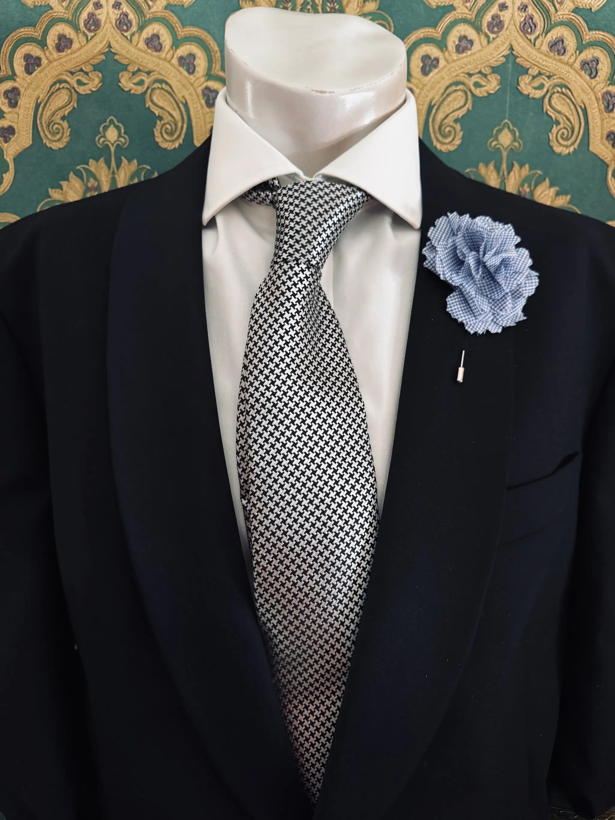 Broochella men's handmade linen carnation brooch in blue check. Pinned to a men's mannequin wearing a black suit and a tie. The background is green and gold damask wallpaper.