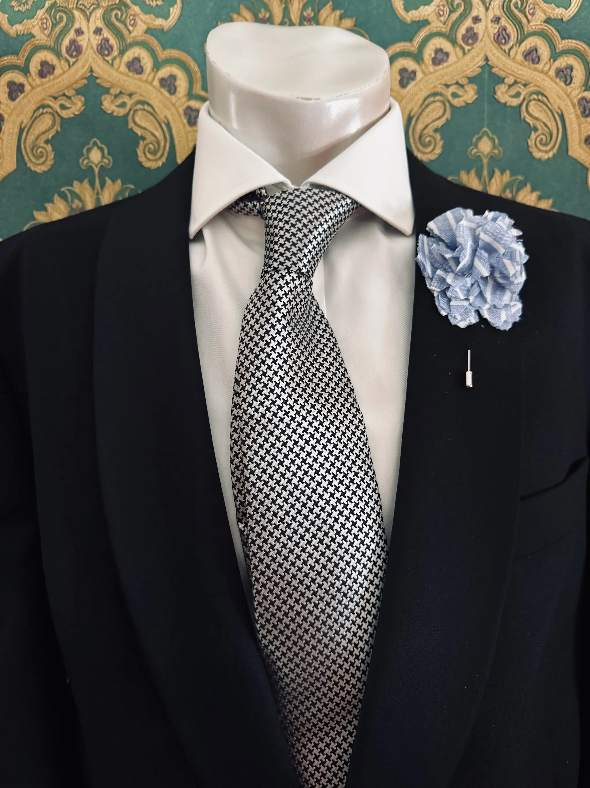 Broochella men's handmade linen carnation brooch in blue and white stripe. Pinned to a men's mannequin wearing a black suit and a tie. The background is green and gold damask wallpaper.