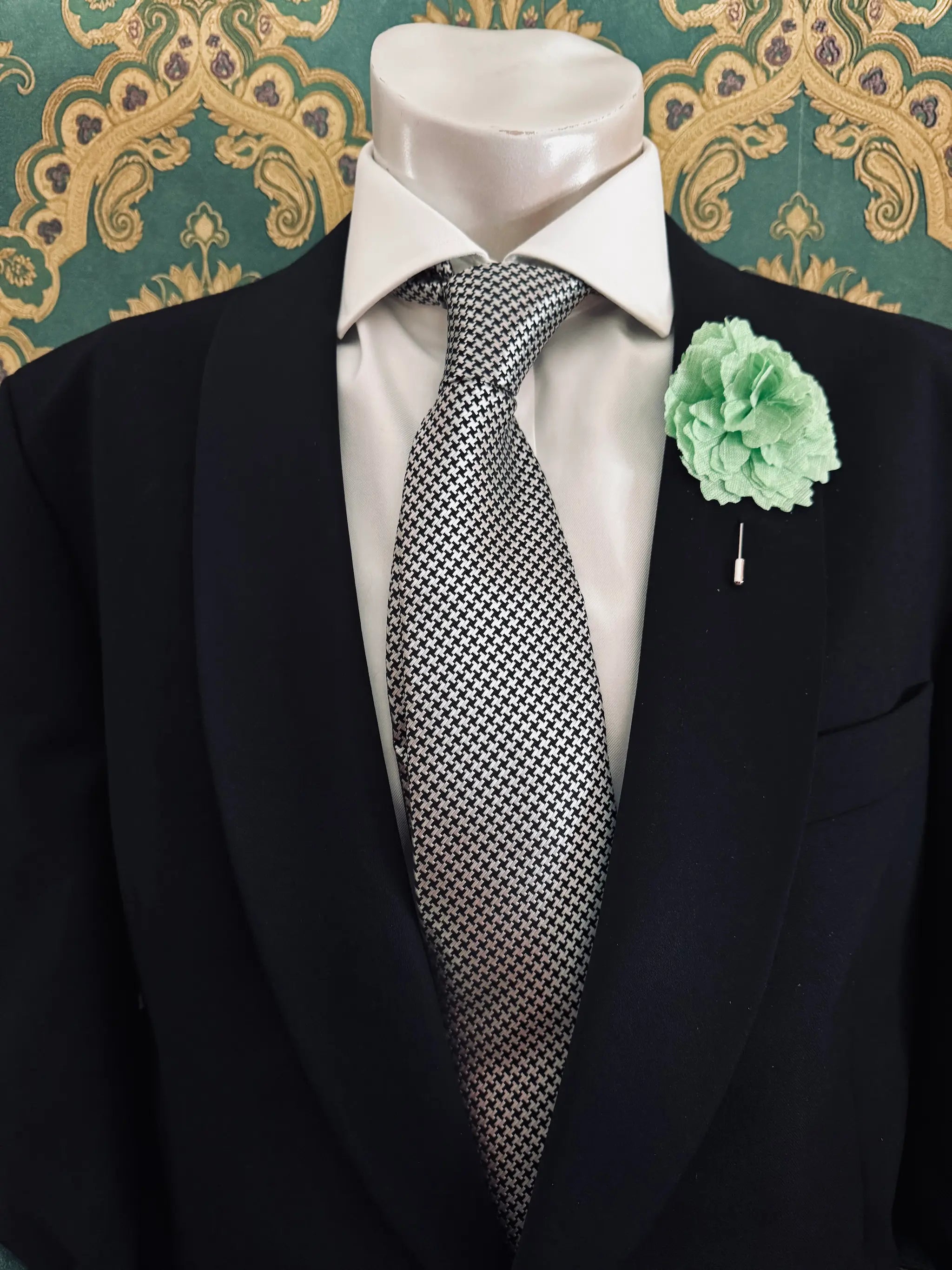 Broochella men's handmade linen carnation brooch in green. Pinned to a men's mannequin wearing a black suit and a tie. The background is green and gold damask wallpaper.