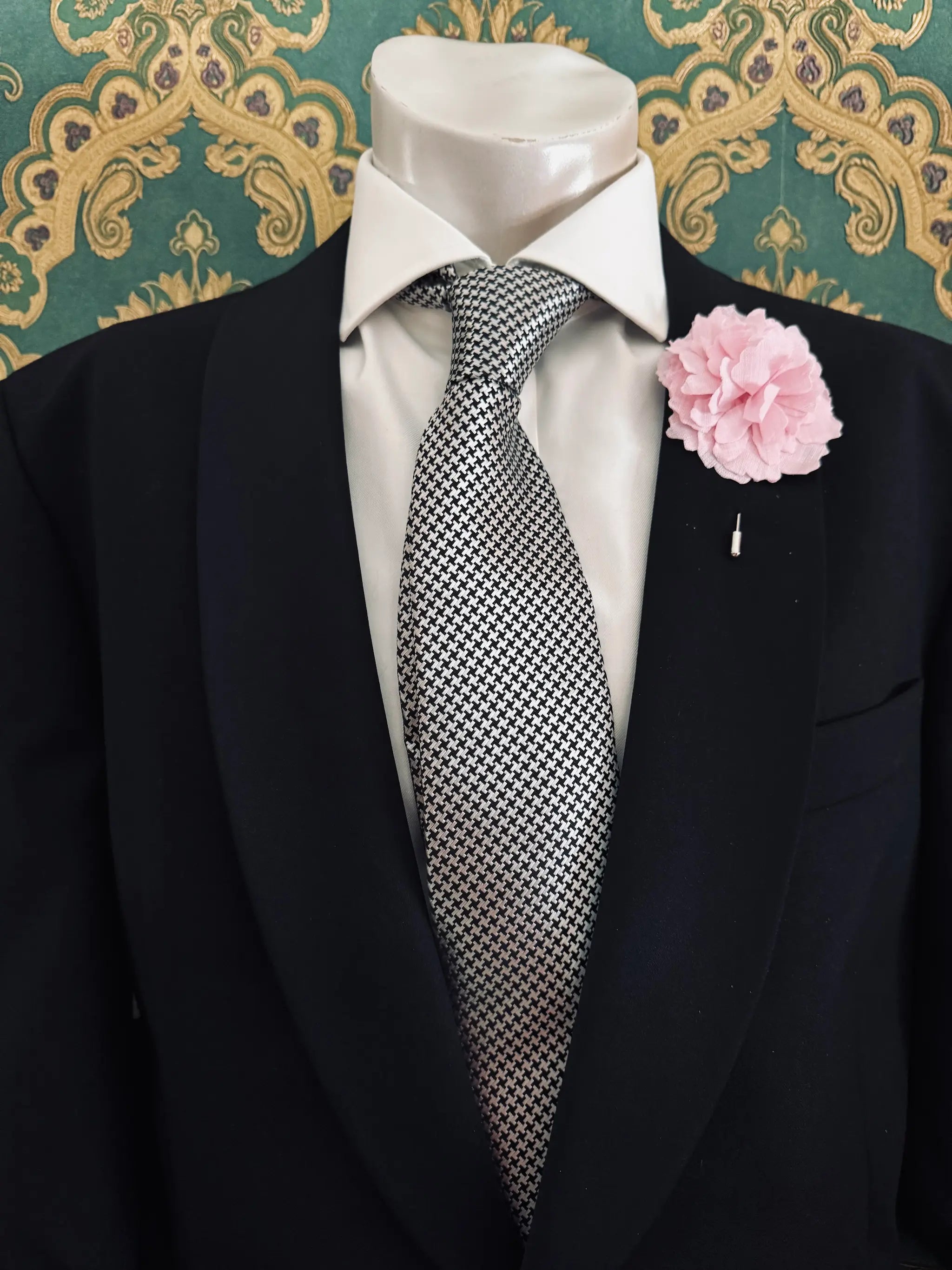 Broochella men's handmade linen carnation brooch in pink. Pinned to a men's mannequin wearing a black suit and a tie. The background is green and gold damask wallpaper.