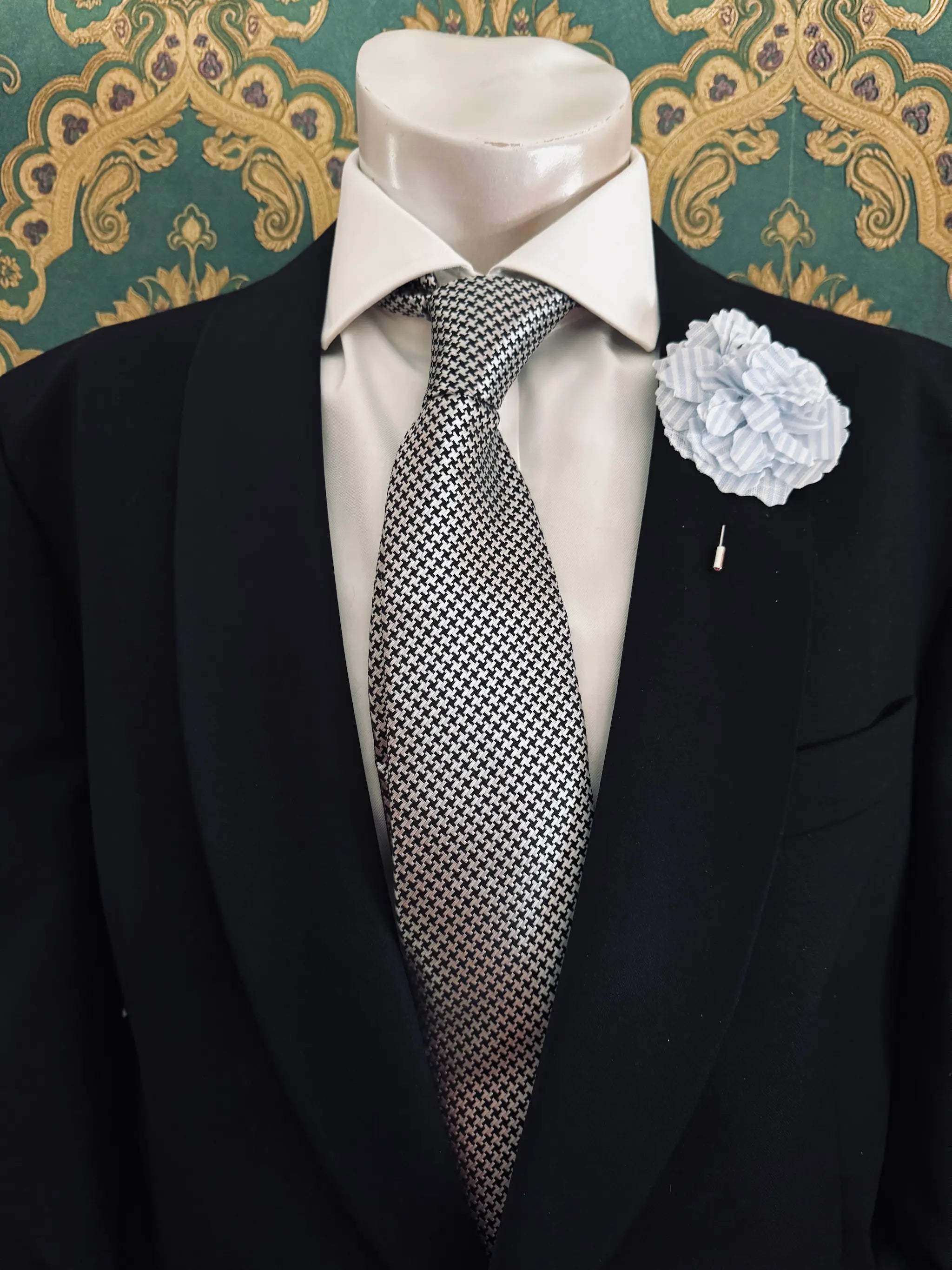 Broochella men's handmade linen carnation brooch in blue and white stripe. Pinned to a men's mannequin wearing a black suit and a tie. The background is green and gold damask wallpaper.