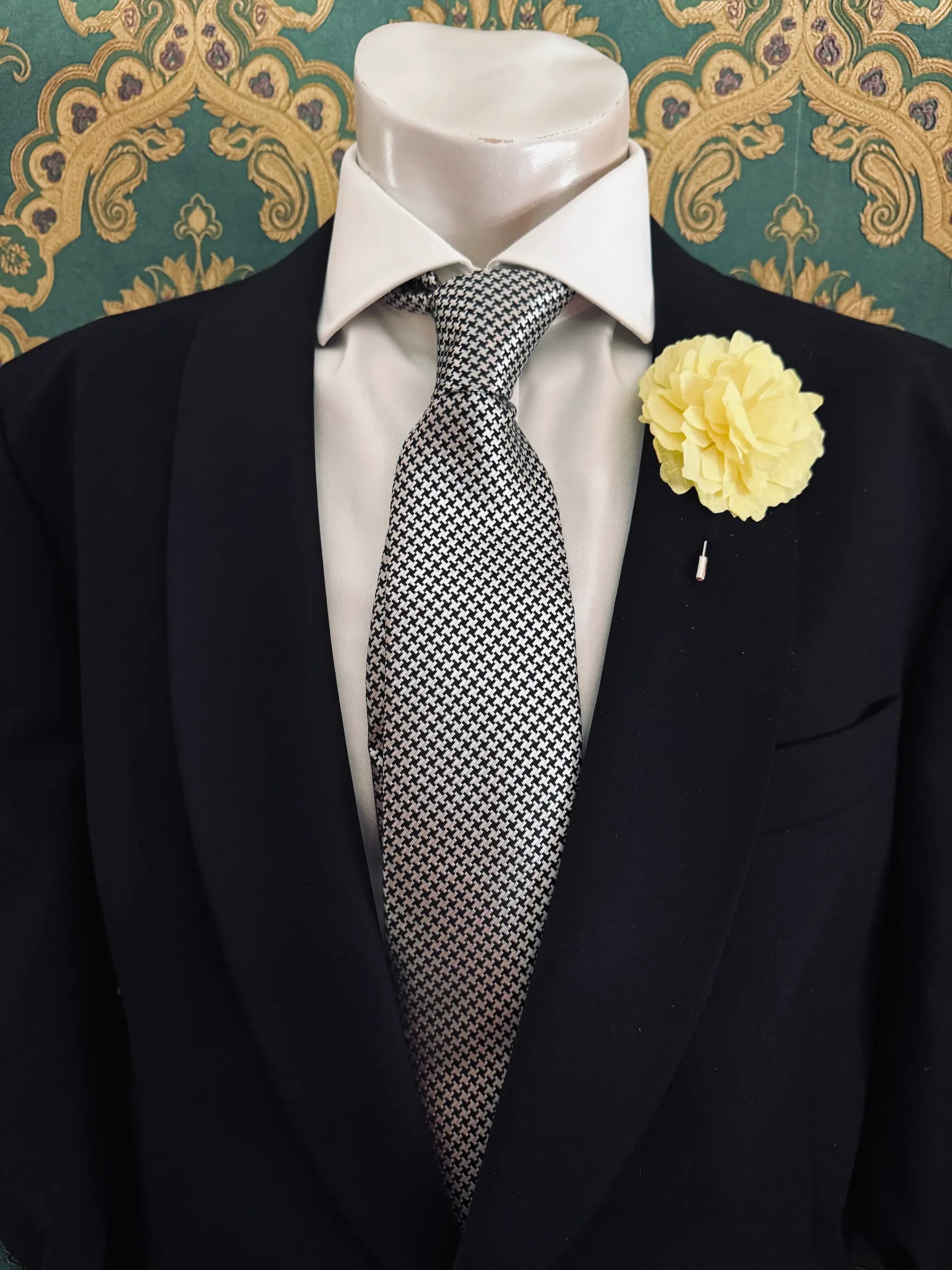 Broochella men's handmade linen carnation brooch in yellow. Pinned to a men's mannequin wearing a black suit and a tie. The background is green and gold damask wallpaper.