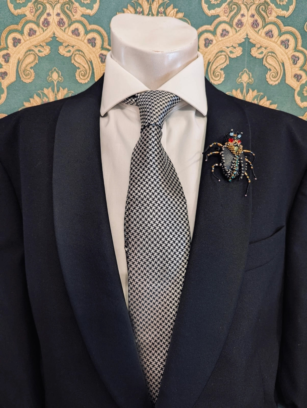 Broochella men's hand beaded cockroach brooch in black leather, with multi coloured beaded head and legs. Pinned to a men's mannequin wearing a black suit and a tie. The background is green and gold damask wallpaper.