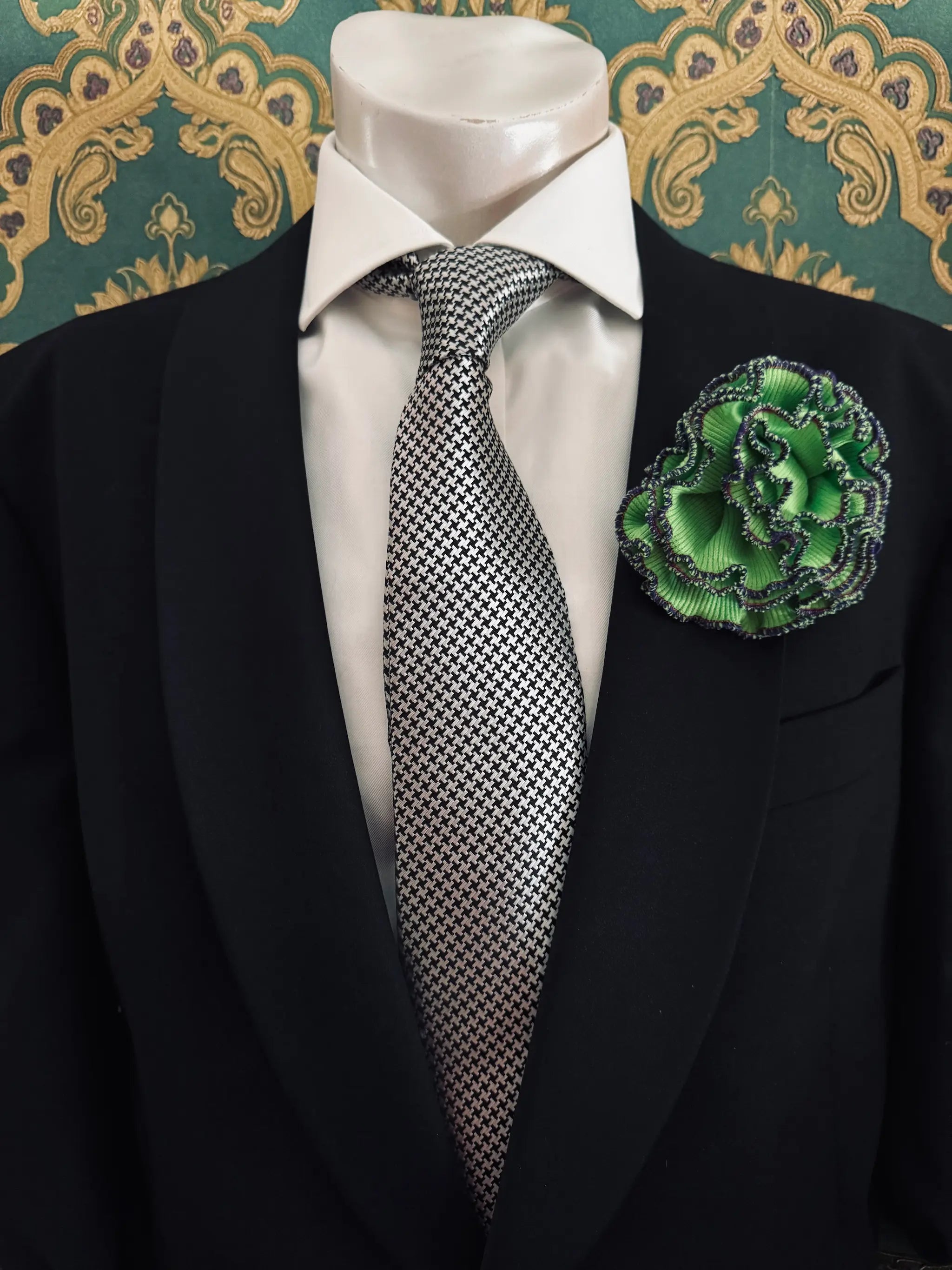 Broochella Men's corallo brooch. Made from green, ribbed, stretch fabric. The edges are finished with purple stitching.  The brooch resembles a bright piece of coral. Pinned to a men's mannequin wearing a black suit and a tie. The background is green and gold damask wallpaper.