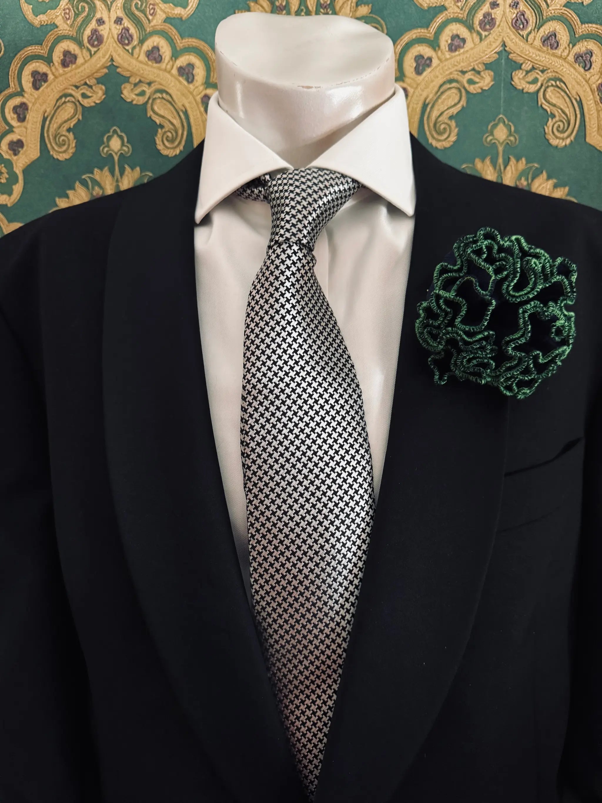 Broochella Men's corallo brooch. Made from navy, ribbed, stretch fabric. The edges are finished with green stitching.  The brooch resembles a bright piece of coral. Pinned to a men's mannequin wearing a black suit and a tie. The background is green and gold damask wallpaper.
