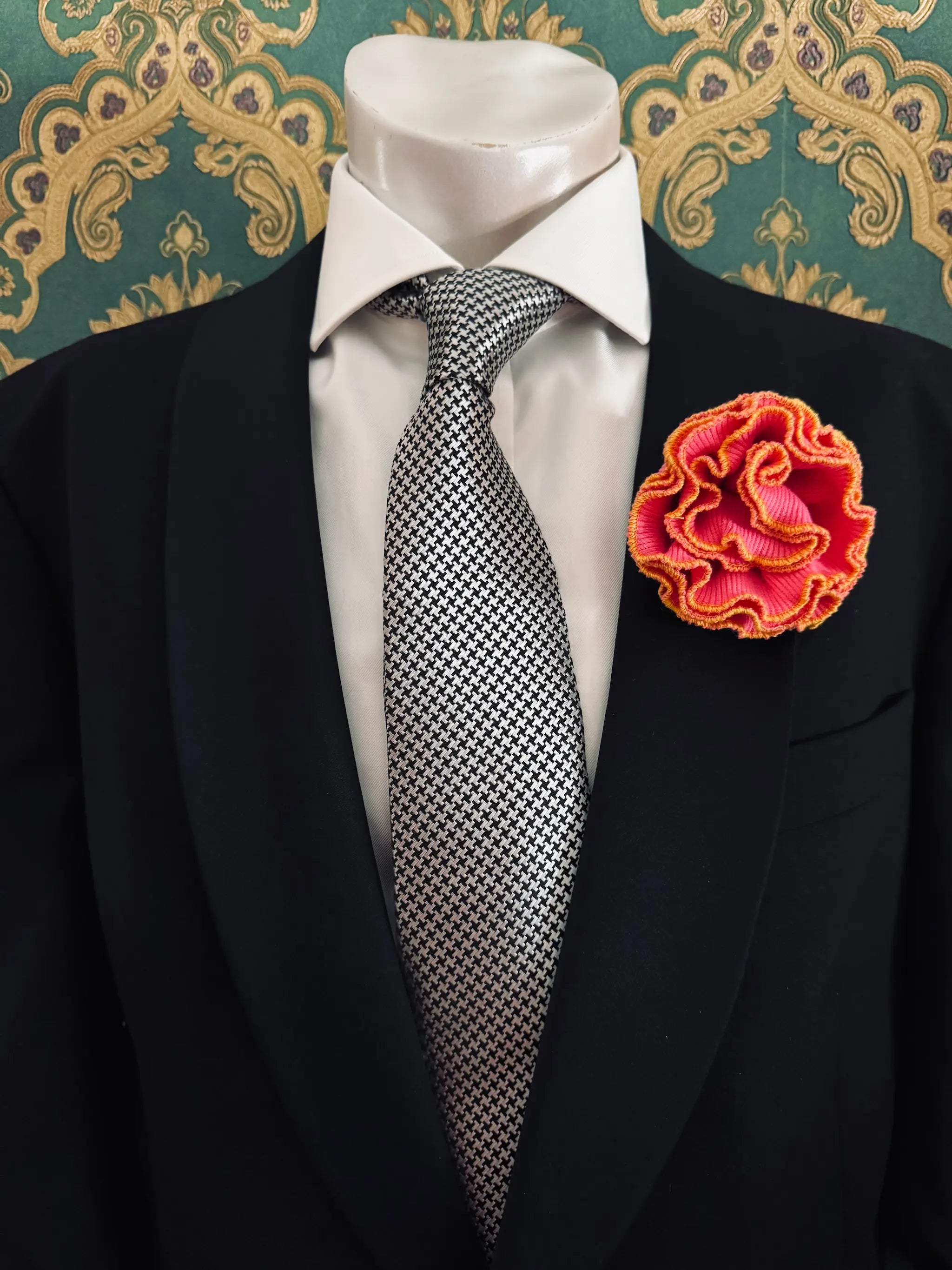 Broochella Men's corallo brooch. Made from pink, ribbed, stretch fabric. The edges are finished with yellow stitching.  The brooch resembles a bright piece of coral. Pinned to a men's mannequin wearing a black suit and a tie. The background is green and gold damask wallpaper.