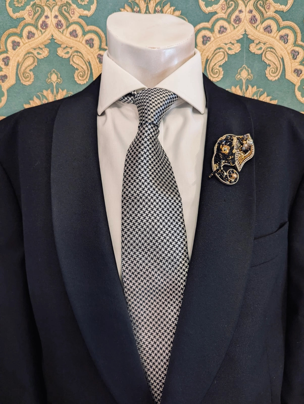 Broochella men's hand beaded elephant brooch in black and gold. Pinned to a men's mannequin wearing a black suit and a tie. The background is green and gold damask wallpaper.