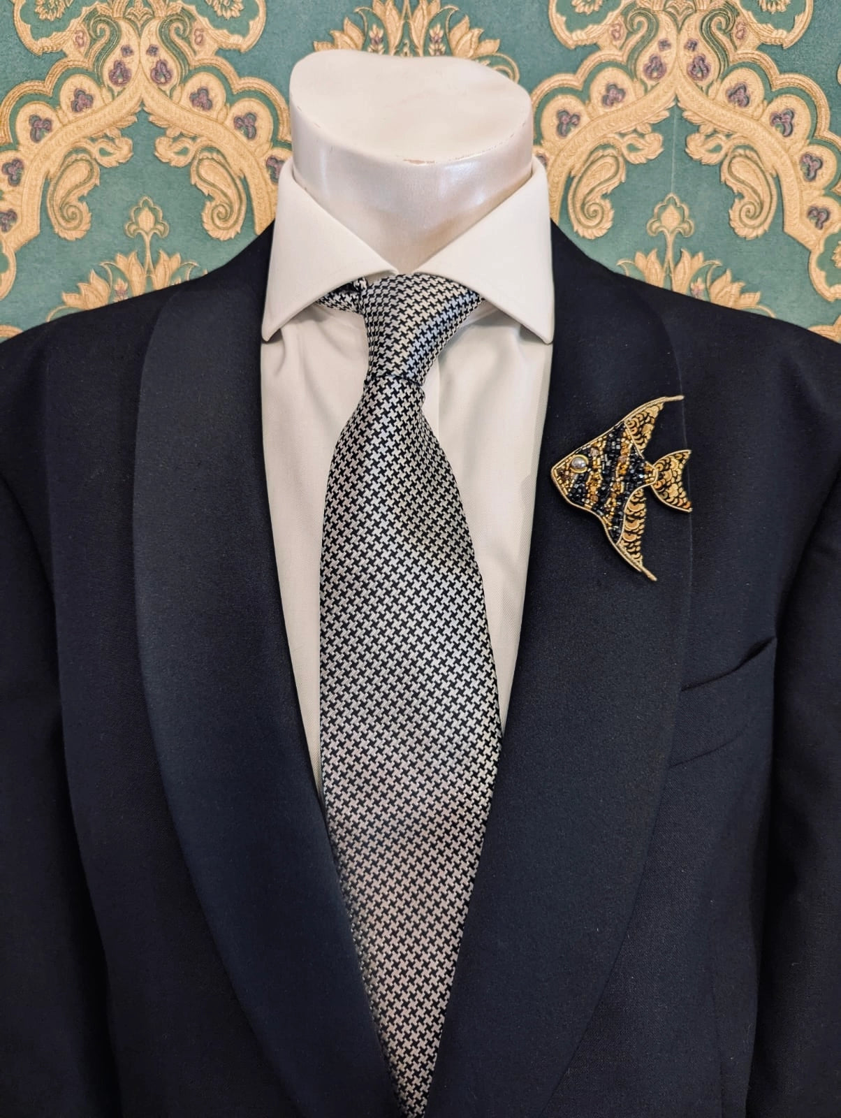 Broochella men's hand beaded fish brooch in black and gold. Pinned to a men's mannequin wearing a black suit and a tie. The background is green and gold damask wallpaper.