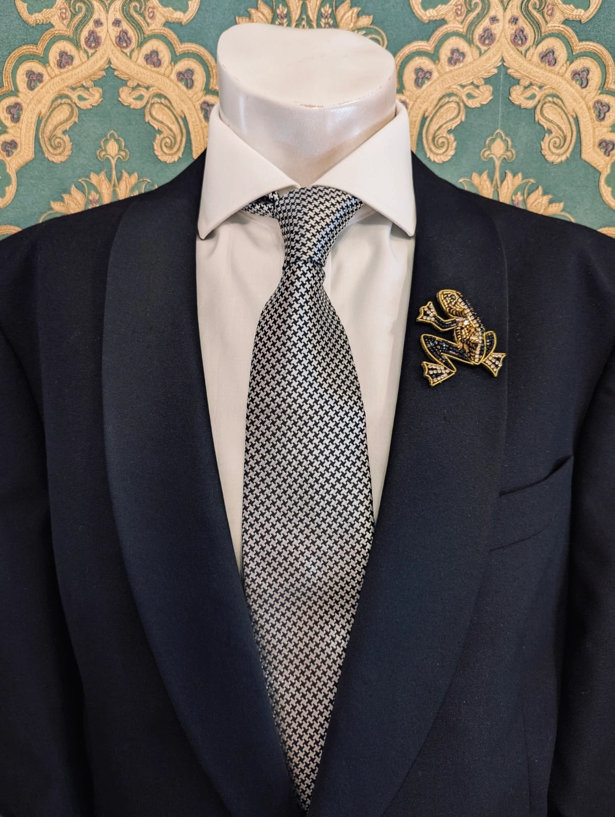 Broochella men's hand beaded frog brooch in black and gold. Pinned to a men's mannequin wearing a black suit and a tie. The background is green and gold damask wallpaper.