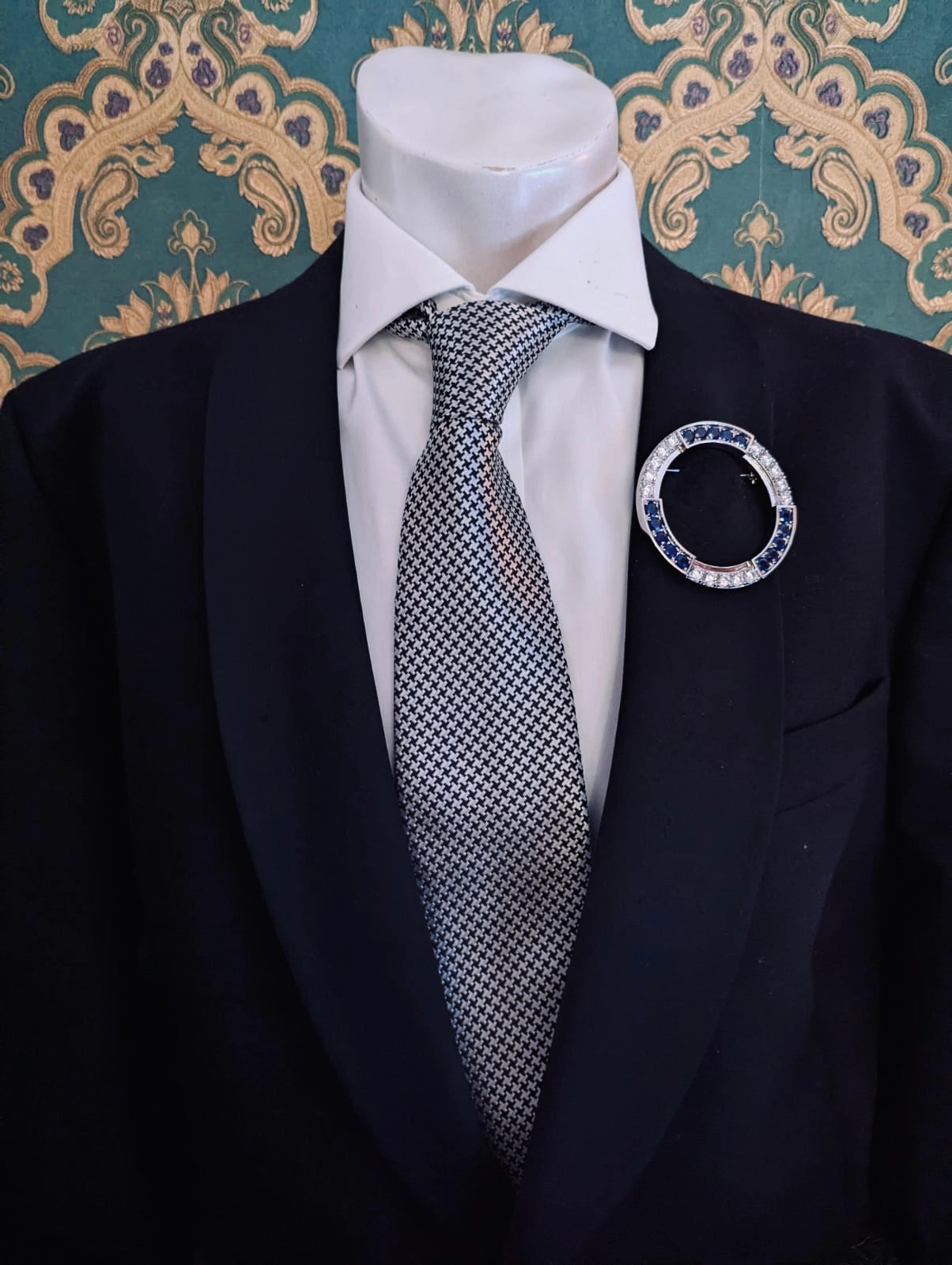 Broochella full moon brooch. Circular silver metal brooch with blu and clear crystals. Pinned to a men's mannequin wearing a black suit and houndstooth tie. The background is green and gold damask wallpaper.