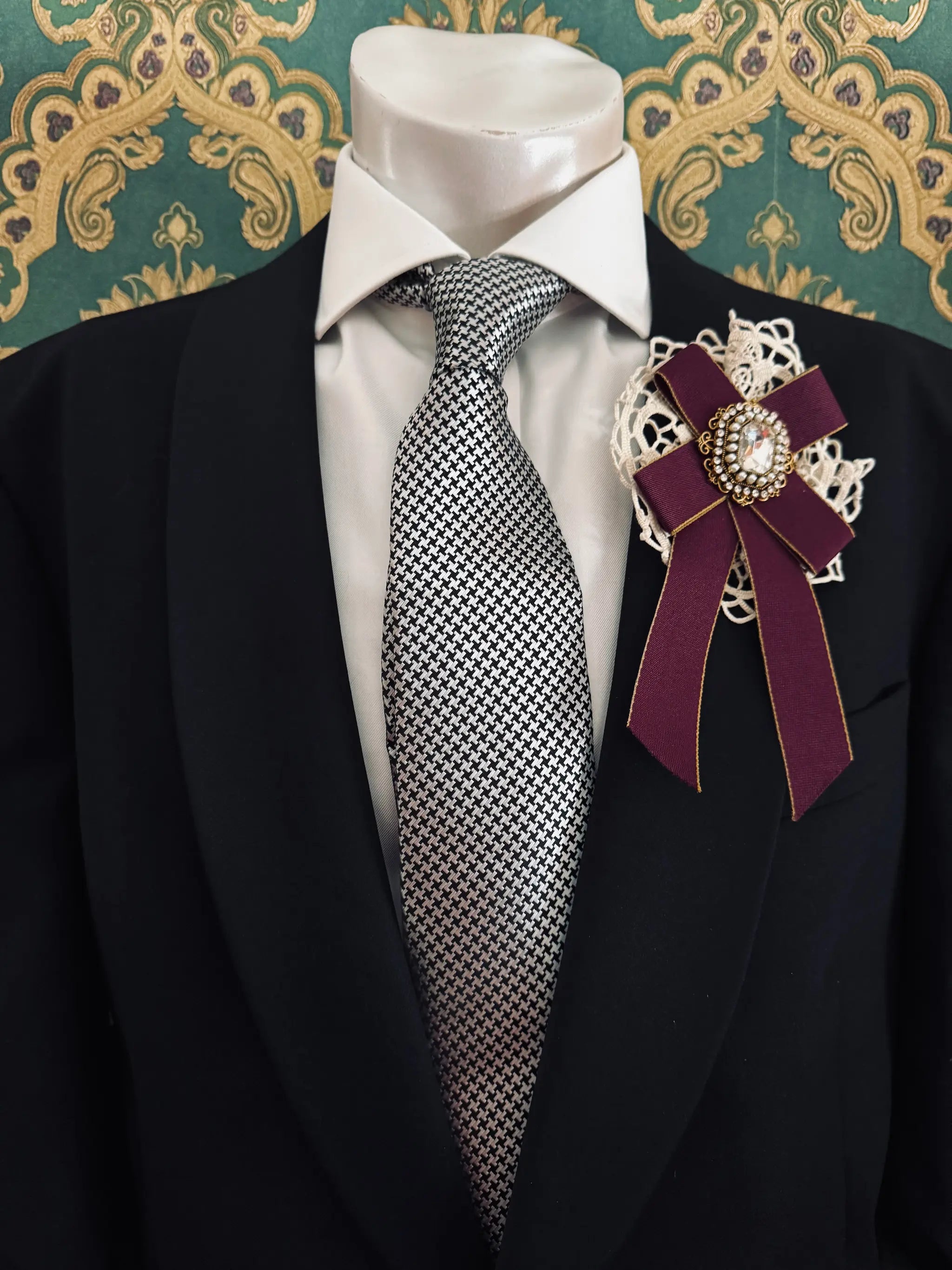 Broochella men's handmade il conte burgindy bow brooch with crochet lace backing and a crystal embellishment centre. Pinned to a men's mannequin wearing a black suit and a tie. The background is green and gold damask wallpaper.