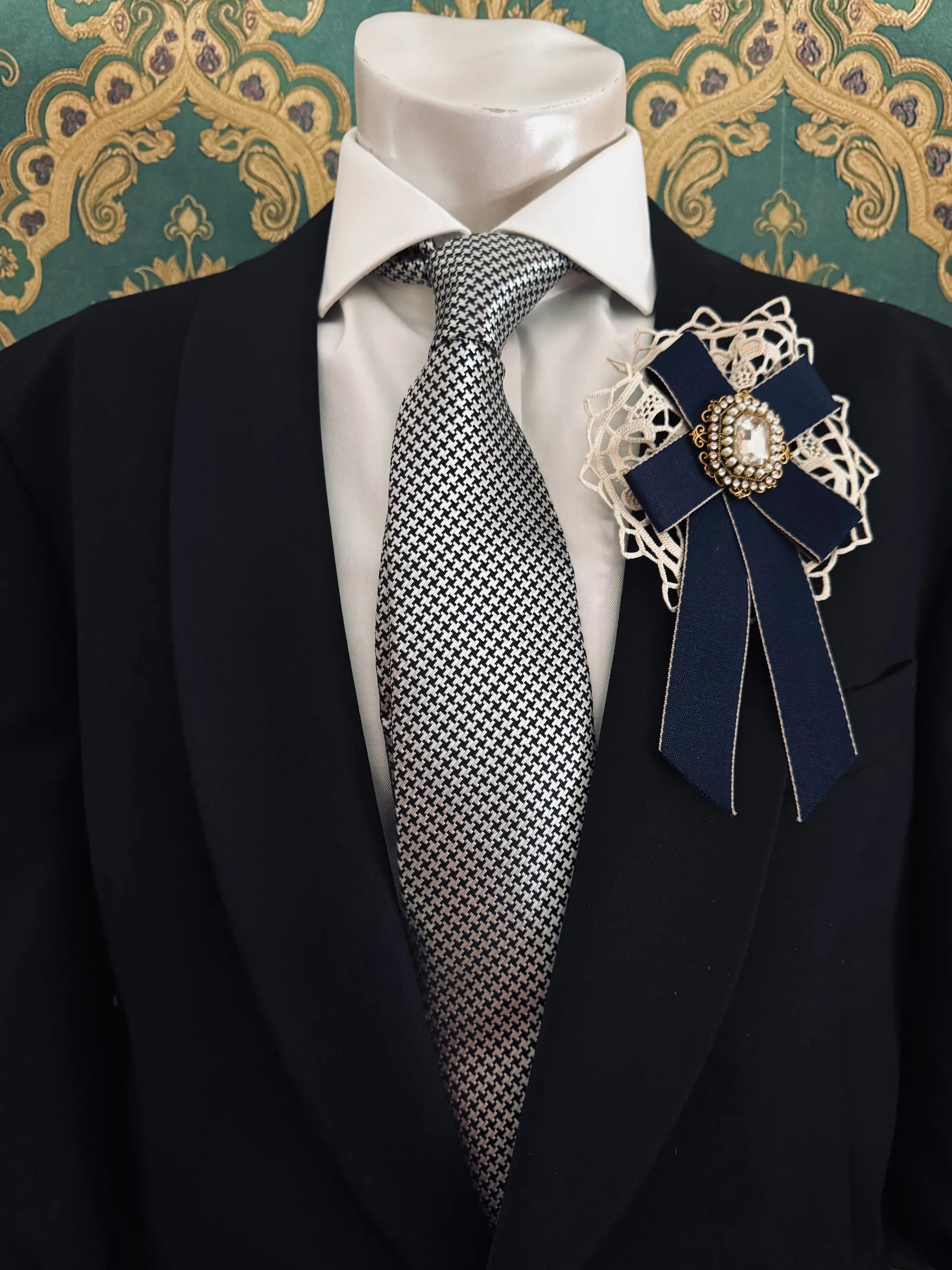 Broochella men's handmade il conte navy bow brooch with crochet lace backing and a crystal embellishment centre. Pinned to a men's mannequin wearing a black suit and a tie. The background is green and gold damask wallpaper.