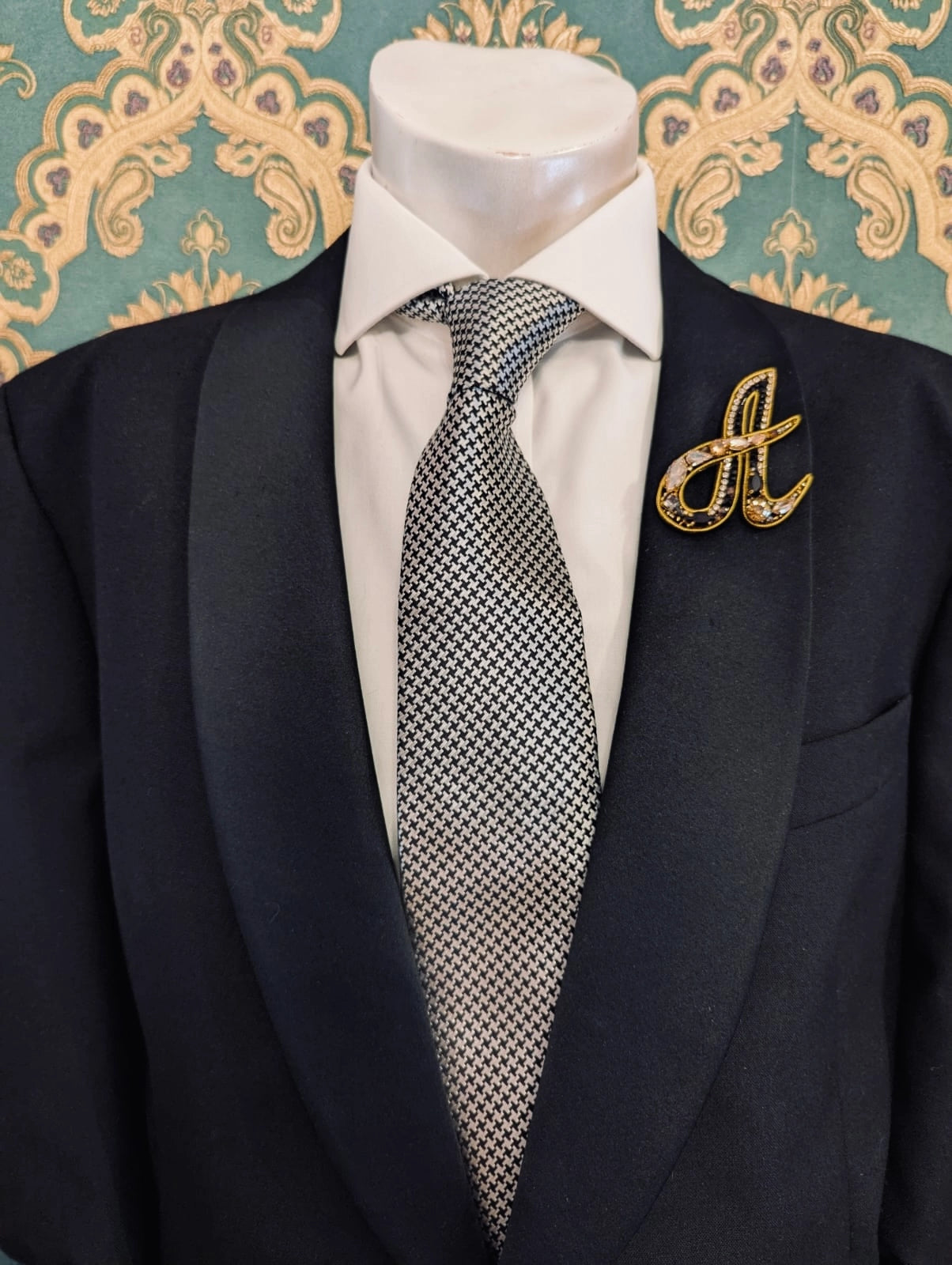 Broochella men's hand beaded A initial brooch in black and gold beads and sequins. Pinned to a men's mannequin wearing a black suit and a tie. The background is green and gold damask wallpaper.
