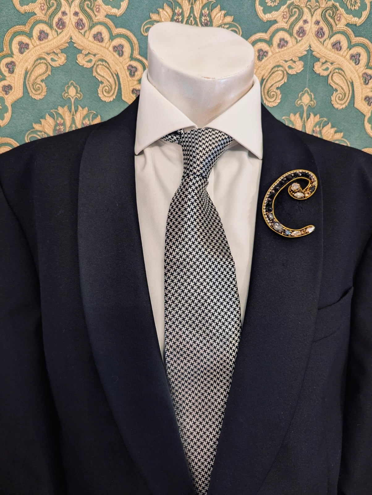 Broochella men's hand beaded C initial brooch in black and gold beads and sequins. Pinned to a men's mannequin wearing a black suit and a tie. The background is green and gold damask wallpaper.