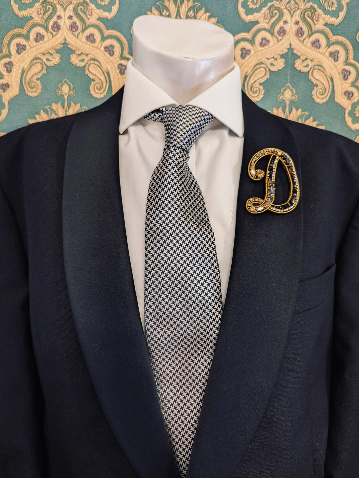 Broochella men's hand beaded D initial brooch in black and gold beads and sequins. Pinned to a men's mannequin wearing a black suit and a tie. The background is green and gold damask wallpaper.