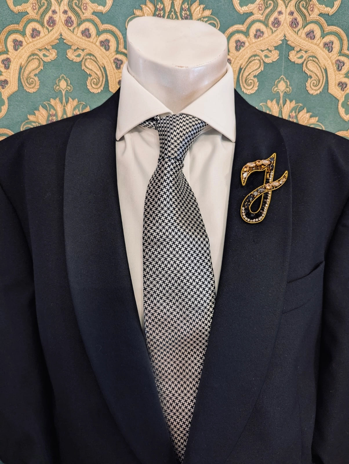 Broochella men's hand beaded I initial brooch in black and gold beads and sequins. Pinned to a men's mannequin wearing a black suit and a tie. The background is green and gold damask wallpaper.