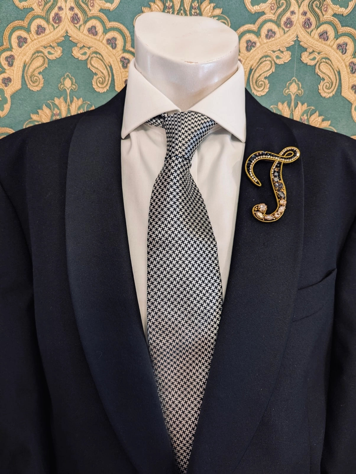 Broochella men's hand beaded J initial brooch in black and gold beads and sequins. Pinned to a men's mannequin wearing a black suit and a tie. The background is green and gold damask wallpaper.