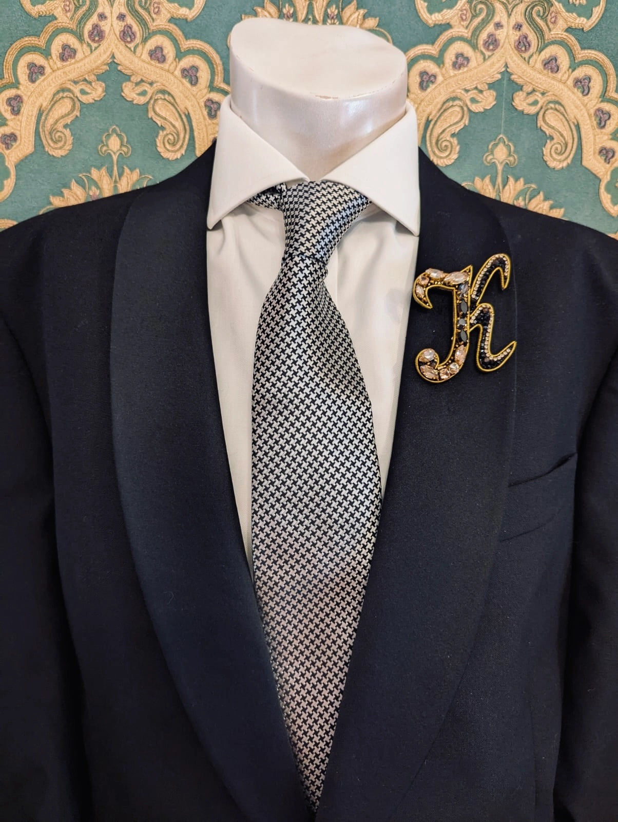 Broochella men's hand beaded K initial brooch in black and gold beads and sequins. Pinned to a men's mannequin wearing a black suit and a tie. The background is green and gold damask wallpaper.