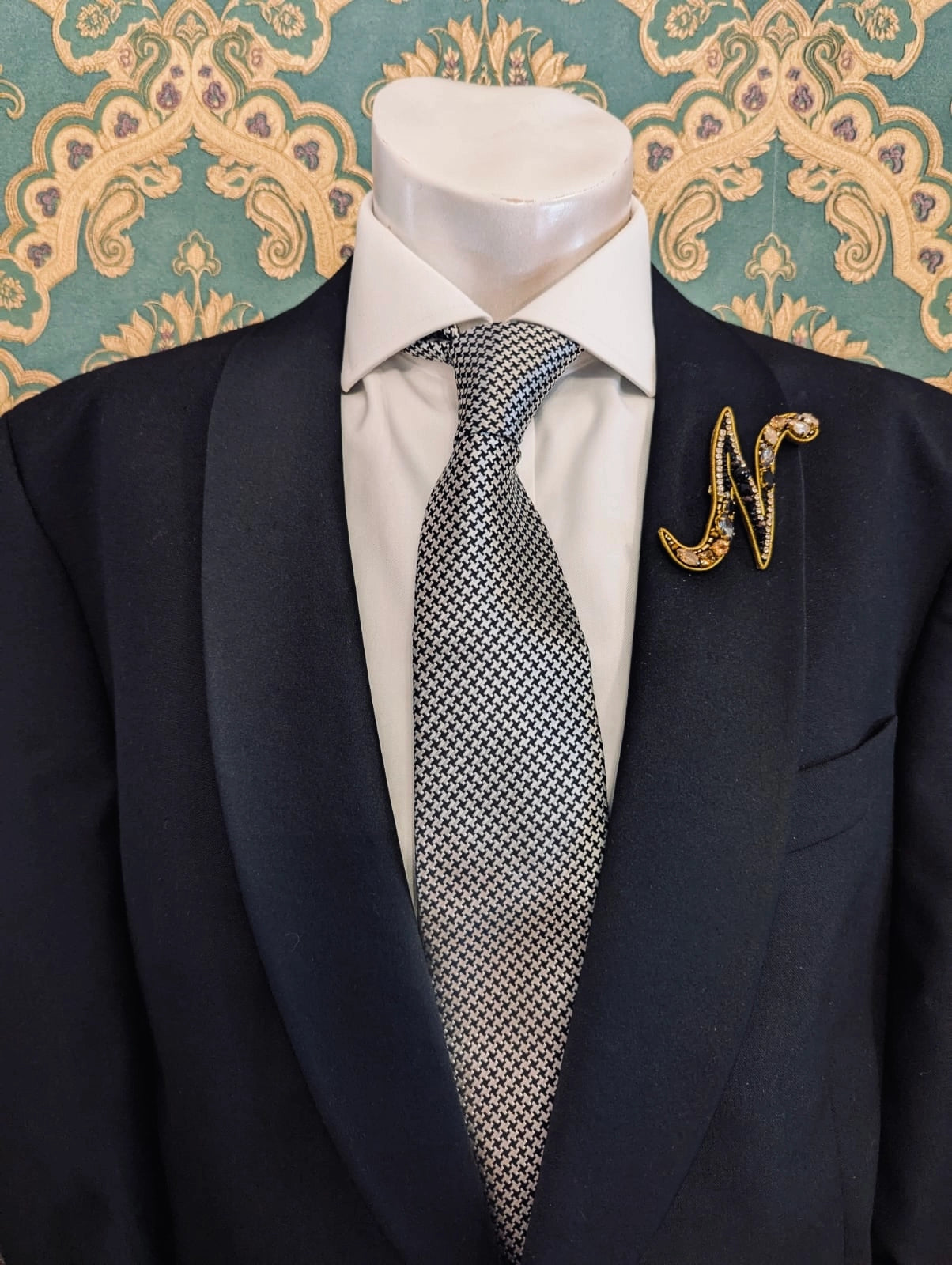 Broochella men's hand beaded N initial brooch in black and gold beads and sequins. Pinned to a men's mannequin wearing a black suit and a tie. The background is green and gold damask wallpaper.