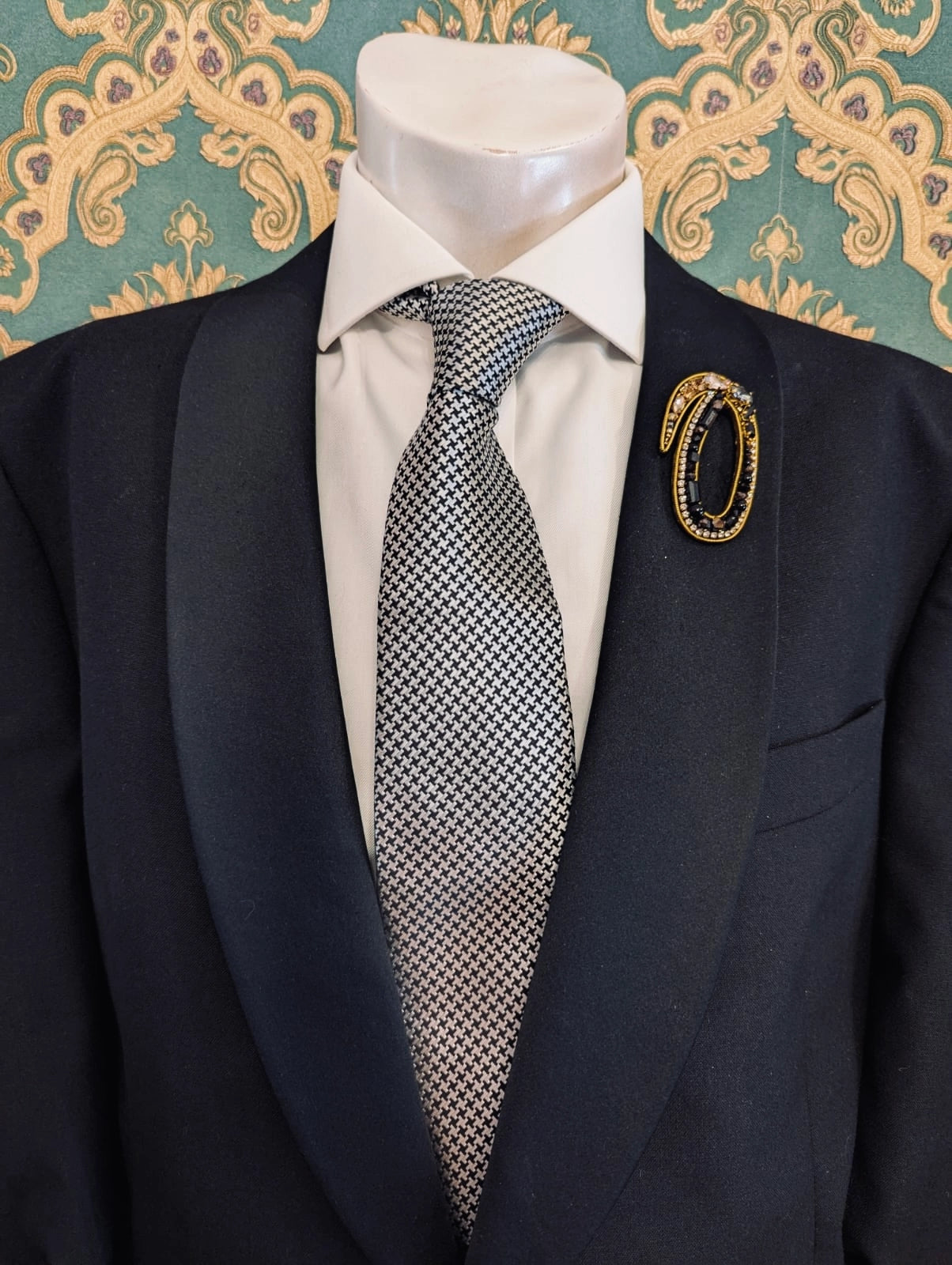 Broochella men's hand beaded O initial brooch in black and gold beads and sequins. Pinned to a men's mannequin wearing a black suit and a tie. The background is green and gold damask wallpaper.
