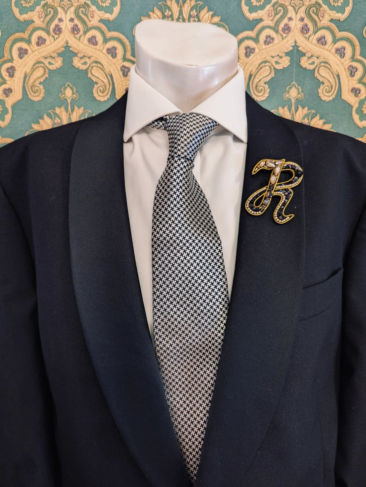 Broochella men's hand beaded R initial brooch in black and gold beads and sequins. Pinned to a men's mannequin wearing a black suit and a tie. The background is green and gold damask wallpaper.
