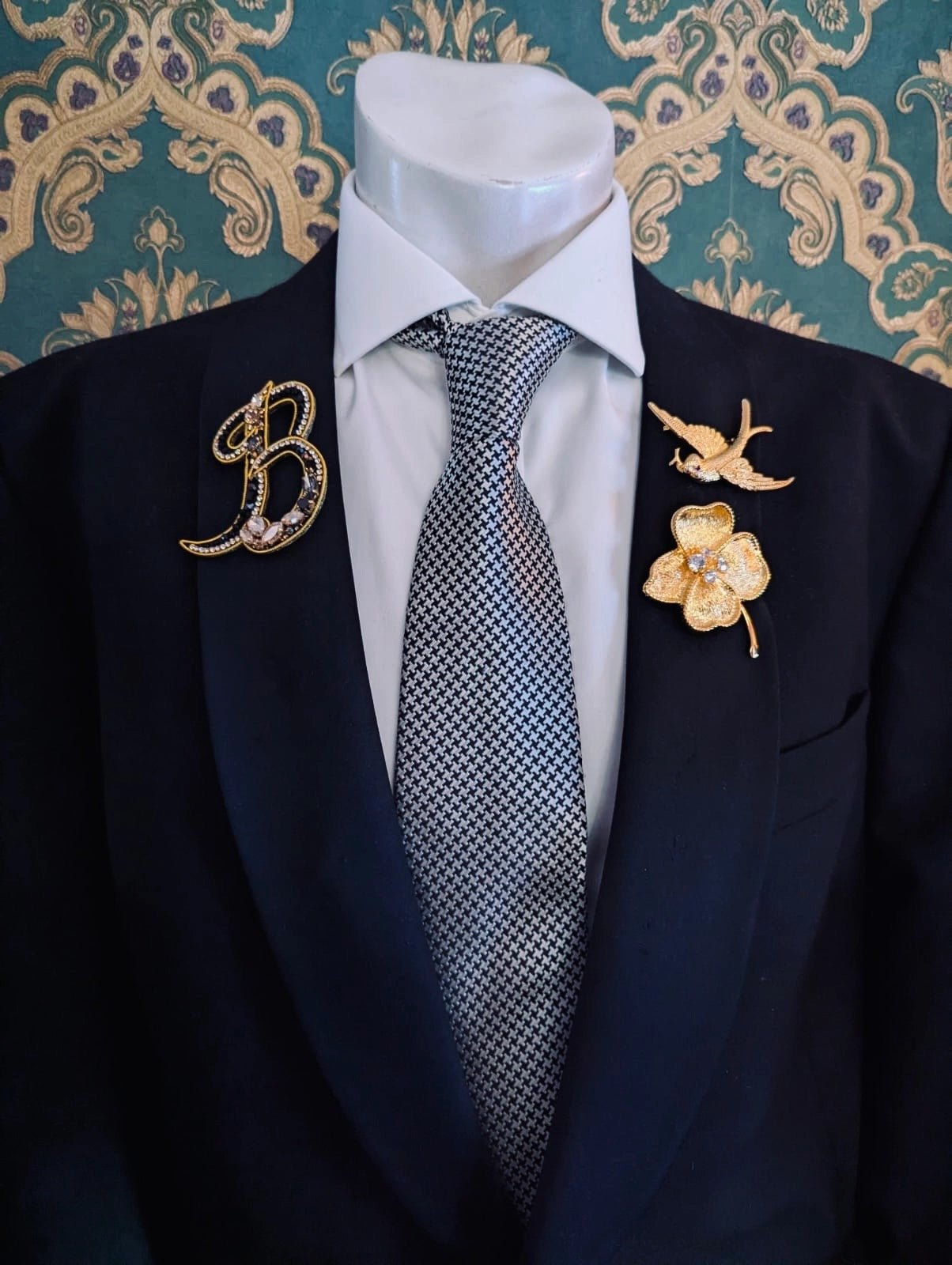 Broochella men's hand beaded B initial brooch in black and gold beads and sequins. Pinned to a men's mannequin wearing a black suit and a tie. The background is green and gold damask wallpaper.