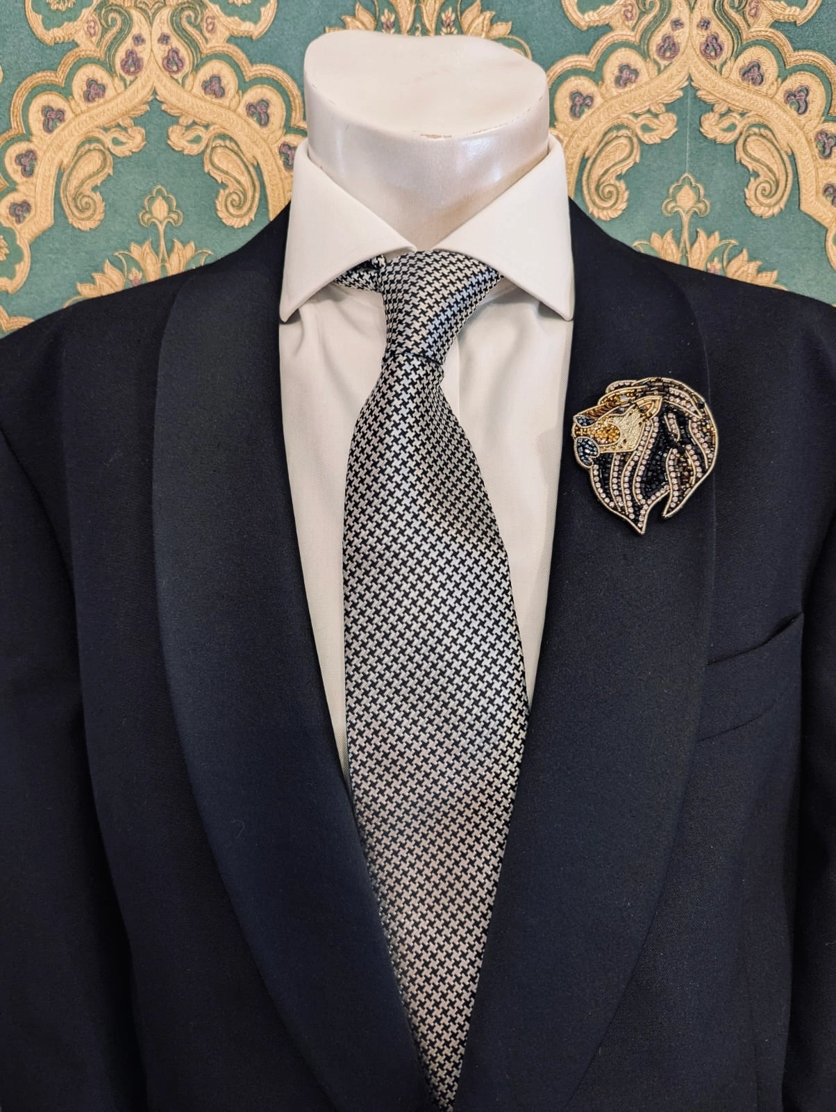 Broochella men's hand beaded lion   brooch in black and gold beads and sequins. Pinned to a men's mannequin wearing a black suit and a tie. The background is green and gold damask wallpaper.