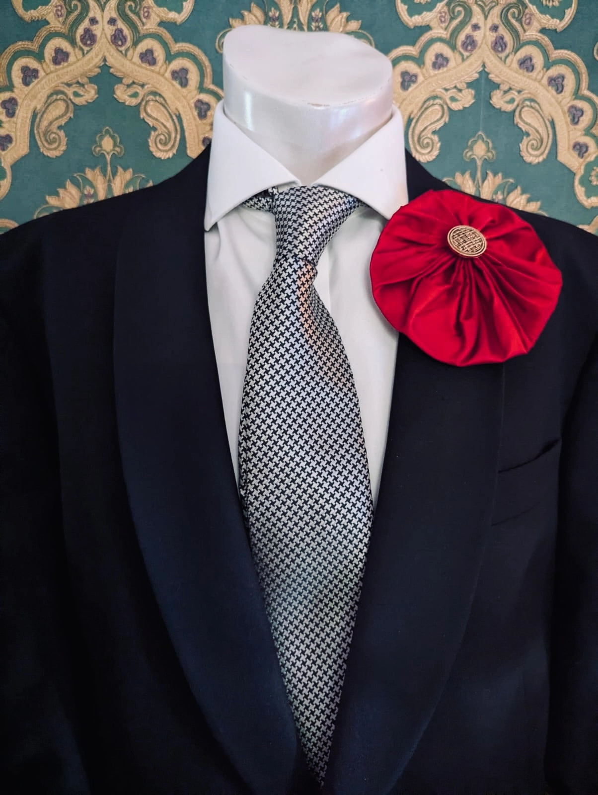 Broochella men's large red handmade silk lollipop brooch with a gold button centre. Pinned to a men's mannequin wearing a black suit and a tie. The background is green and gold damask wallpaper.