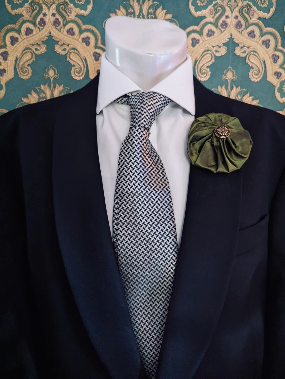 Broochella men's medium green  handmade silk lollipop brooch with a gold button centre. Pinned to a men's mannequin wearing a black suit and a tie. The background is green and gold damask wallpaper.