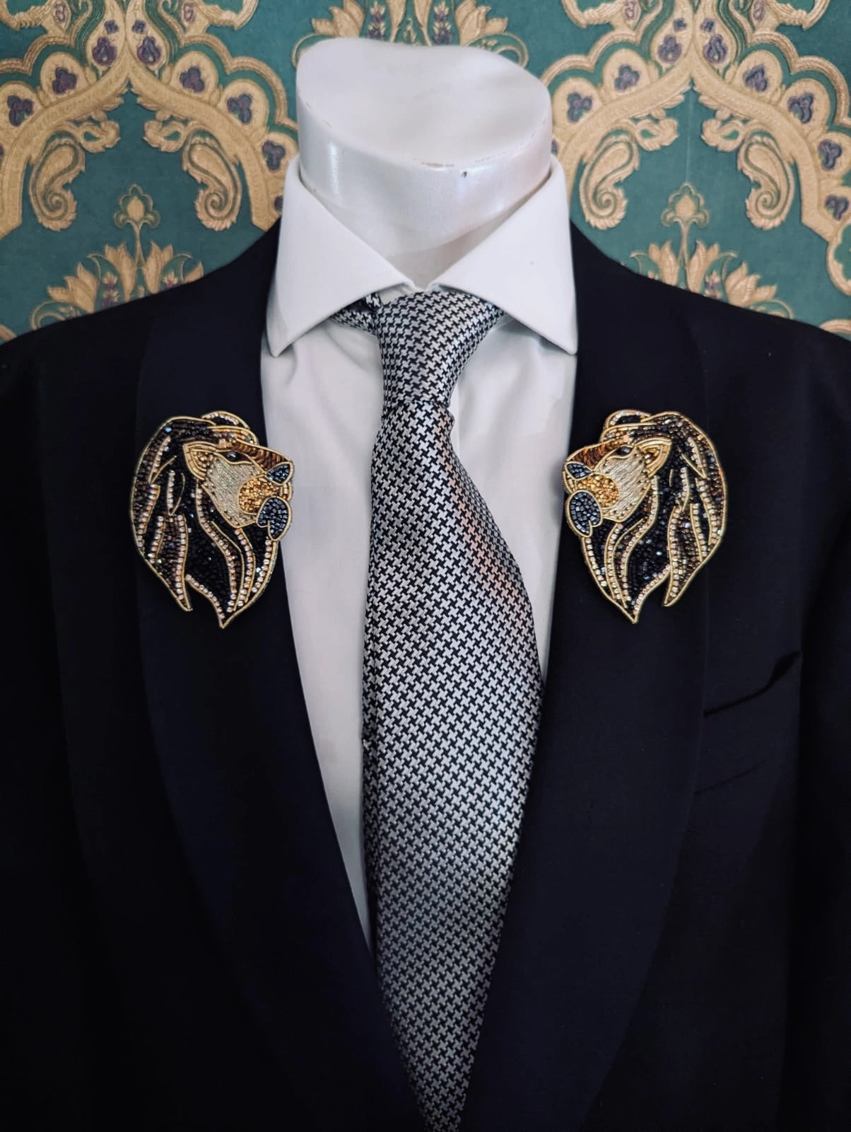 Broochella men's hand beaded mirrored lion head brooches in black and gold beads and sequins. Pinned to a men's mannequin wearing a black suit and a tie. The background is green and gold damask wallpaper.