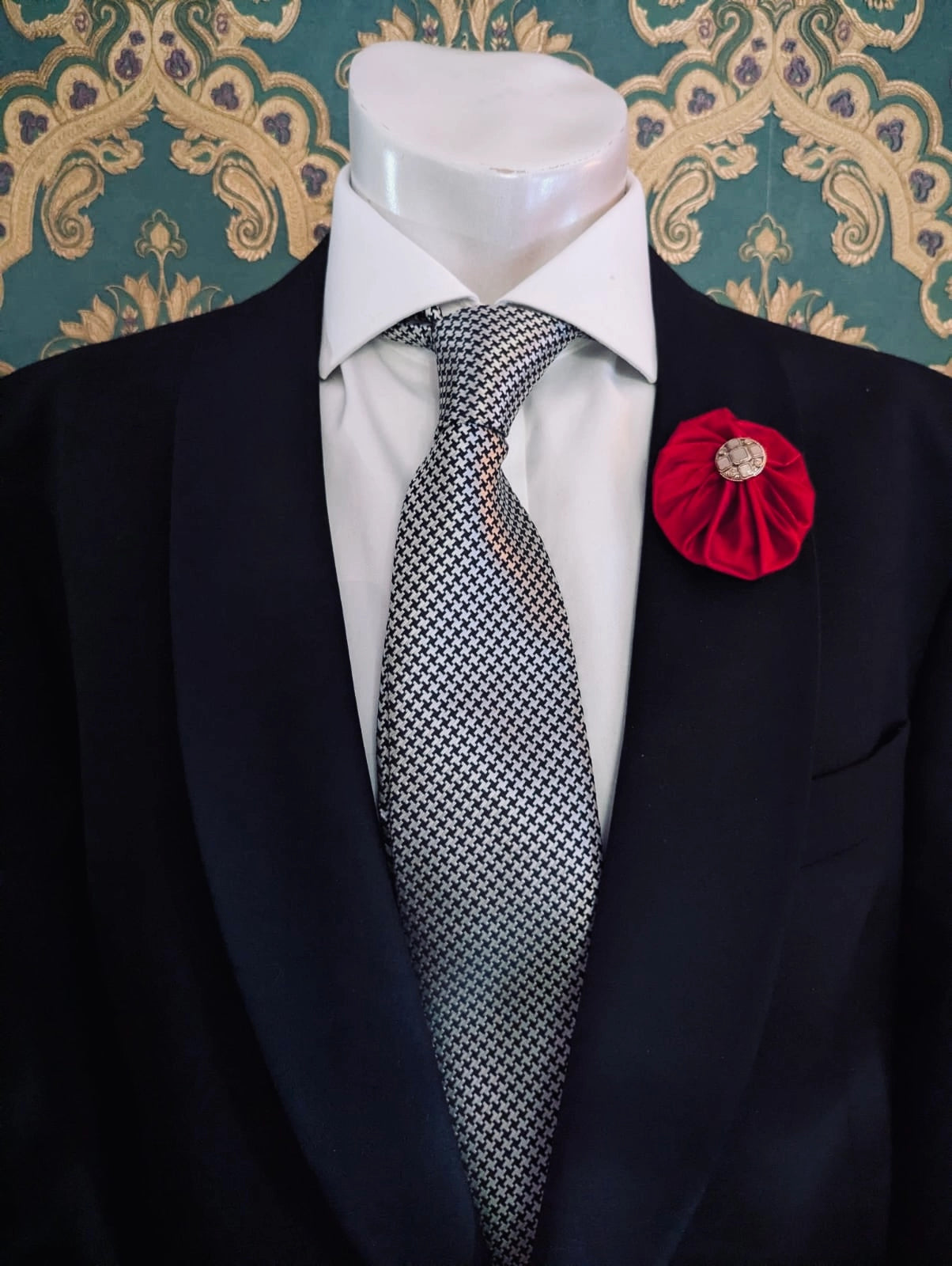 Broochella men's small red handmade silk lollipop brooch with a gold button centre. Pinned to a men's mannequin wearing a black suit and a tie. The background is green and gold damask wallpaper.