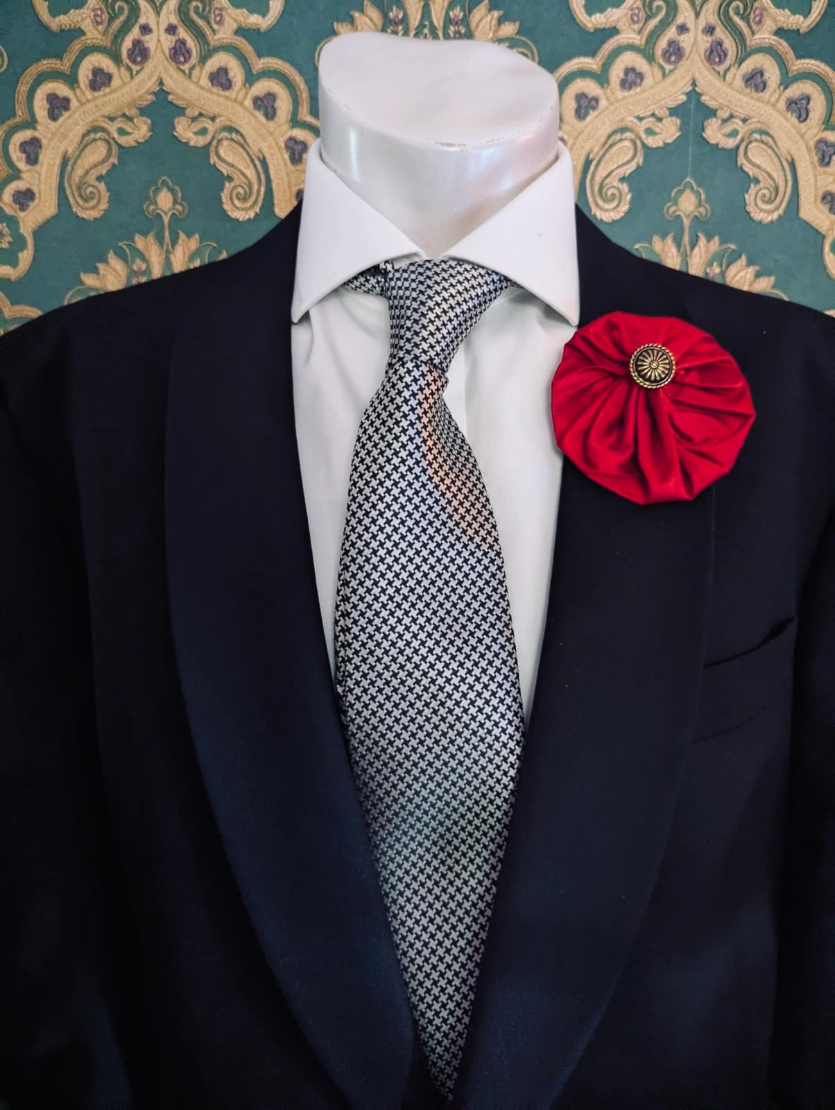 Broochella men's medium red handmade silk lollipop brooch with a gold button centre. Pinned to a men's mannequin wearing a black suit and a tie. The background is green and gold damask wallpaper.