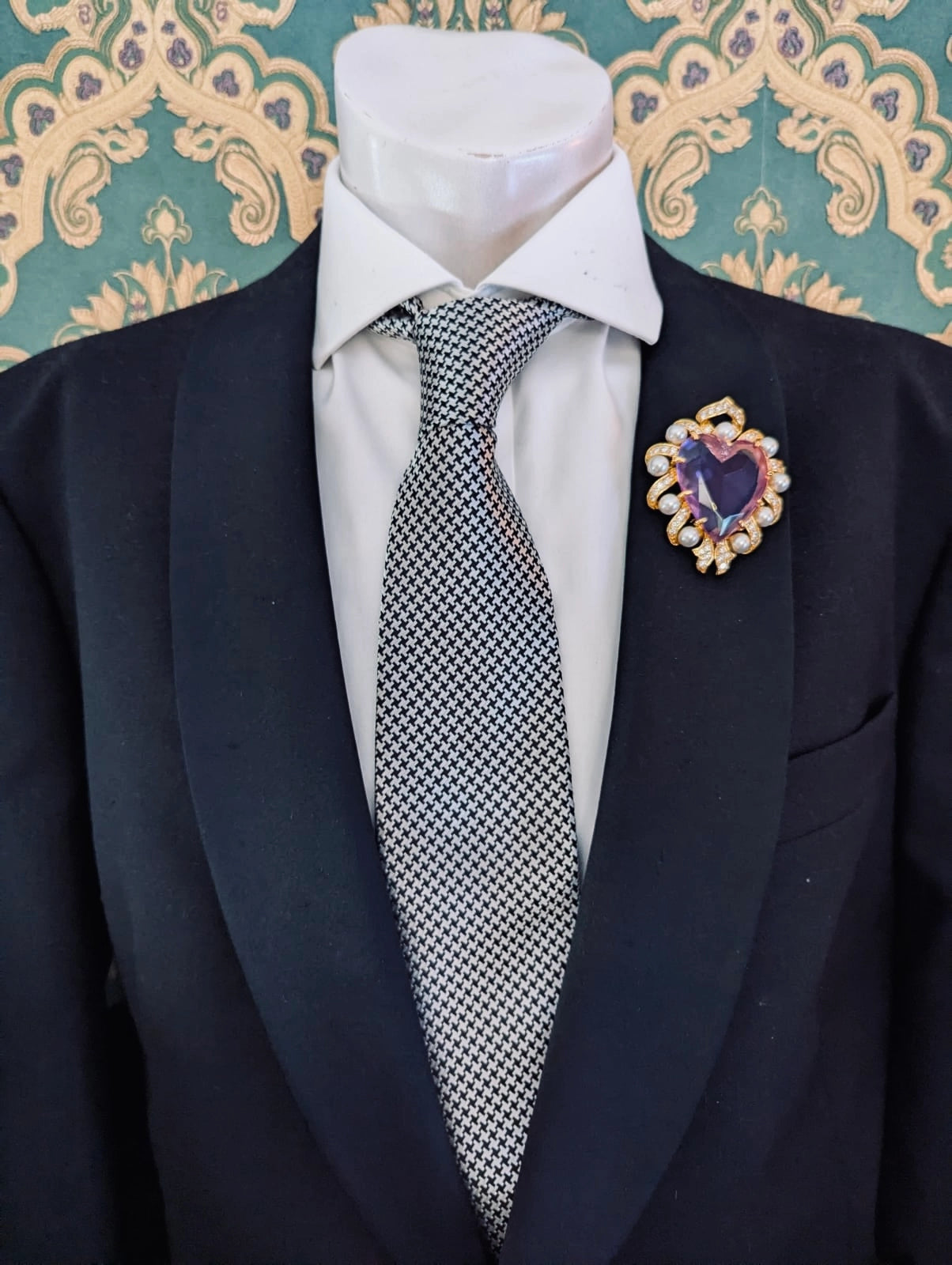 Broochella men's romeo and juliet heart shaped lilac crystal brooch, with gold metal and pearl frame. Pinned to a men's mannequin wearing a black suit and houndstooth tie. The background is green and gold damask wallpaper.