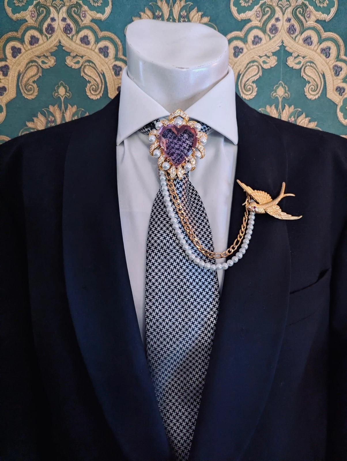 Broochella men's romeo and juliet heart shaped lilac crystal brooch, with gold metal and pearl frame. linked to the broochella swallow brooch with a pearl and gold chain. Pinned to a men's mannequin wearing a black suit and houndstooth tie. The background is green and gold damask wallpaper.