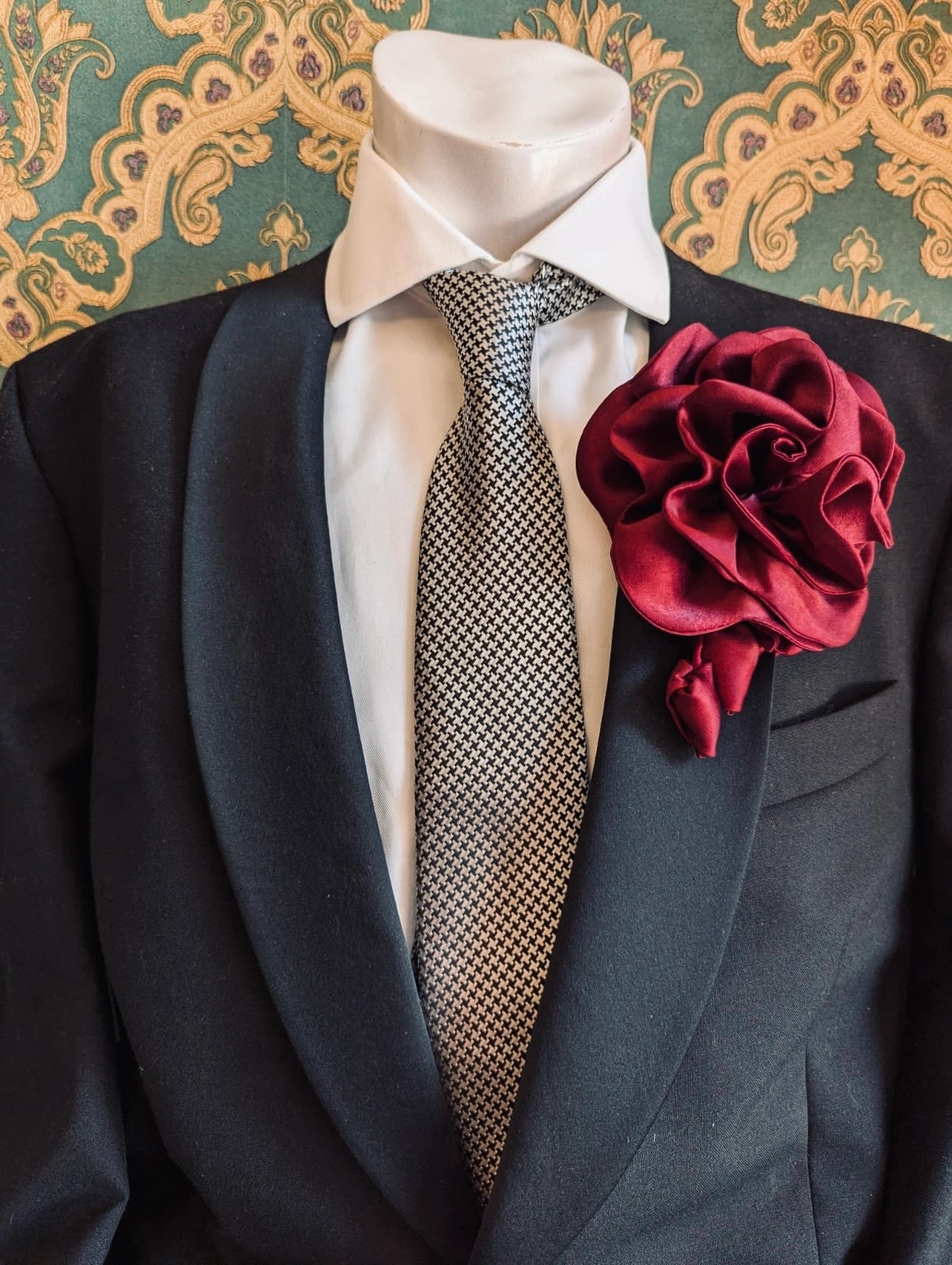 Broochella men's rosetta handmade, designer, satin flower brooch in burgundy. Pinned on a men's mannequin wearing a black suit and a houndstooth tie. The background is green and gold damask wallpaper. 