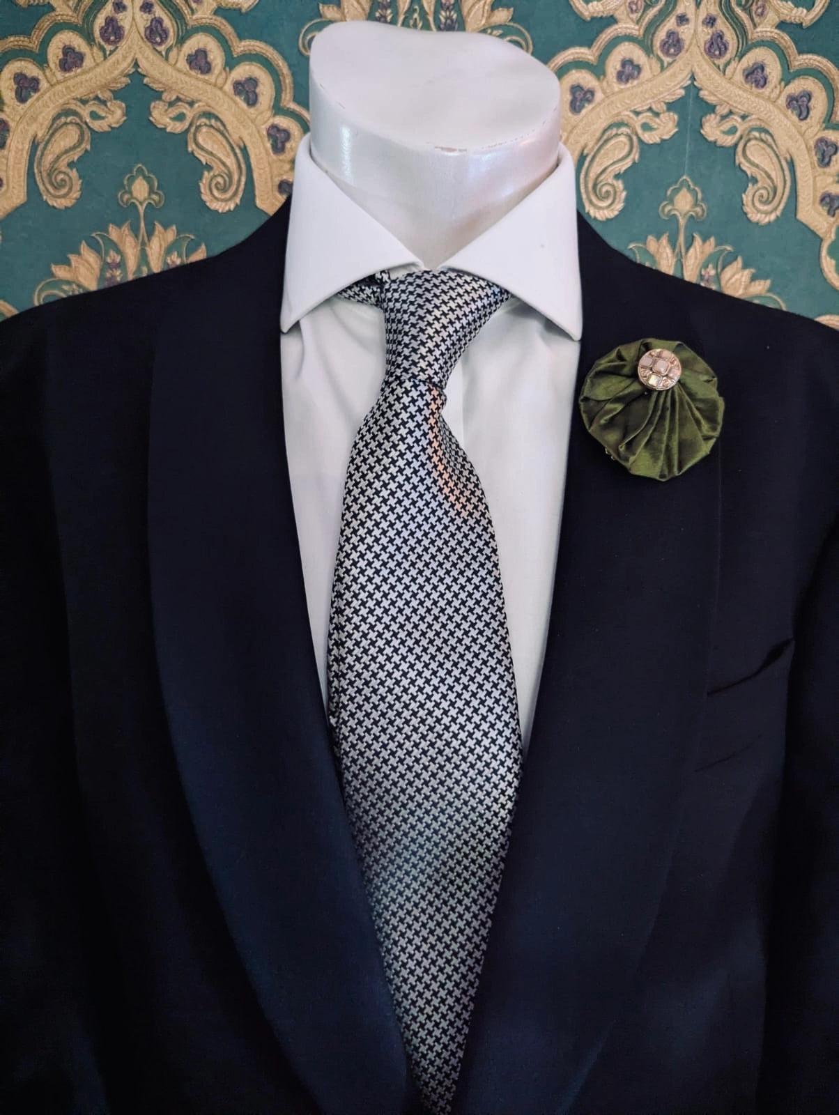 Broochella men's small green handmade silk lollipop brooch with a gold button centre. Pinned to a men's mannequin wearing a black suit and a tie. The background is green and gold damask wallpaper.