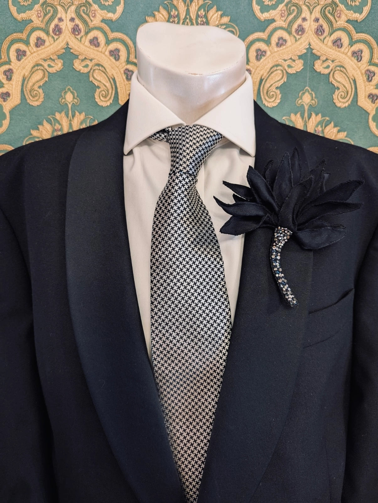 Broochella men's large tulipano hand beaded black flower brooch, with vrystal stem. Pinned to a men's mannequin wearing a black suit and a tie. The background is green and gold damask wallpaper.