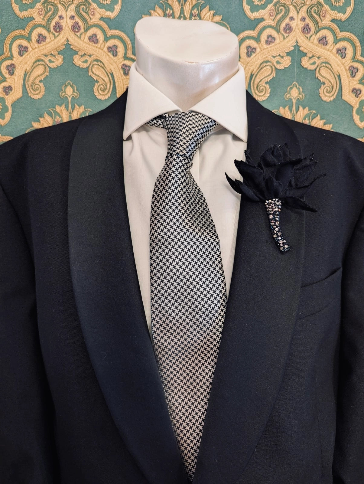 Broochella men's small tulipano hand beaded black flower brooch, with vrystal stem. Pinned to a men's mannequin wearing a black suit and a tie. The background is green and gold damask wallpaper.