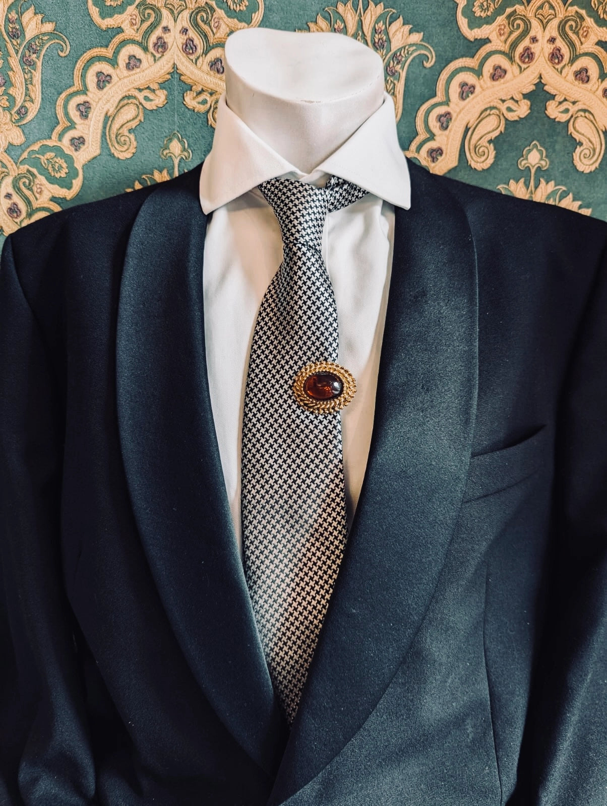 Broochella vintage amber crest tie clip. The centre is a amber stone, the outer is gold metal. Pinned on a men's mannequin in a black suit. The background is green and gold damask wallapaper