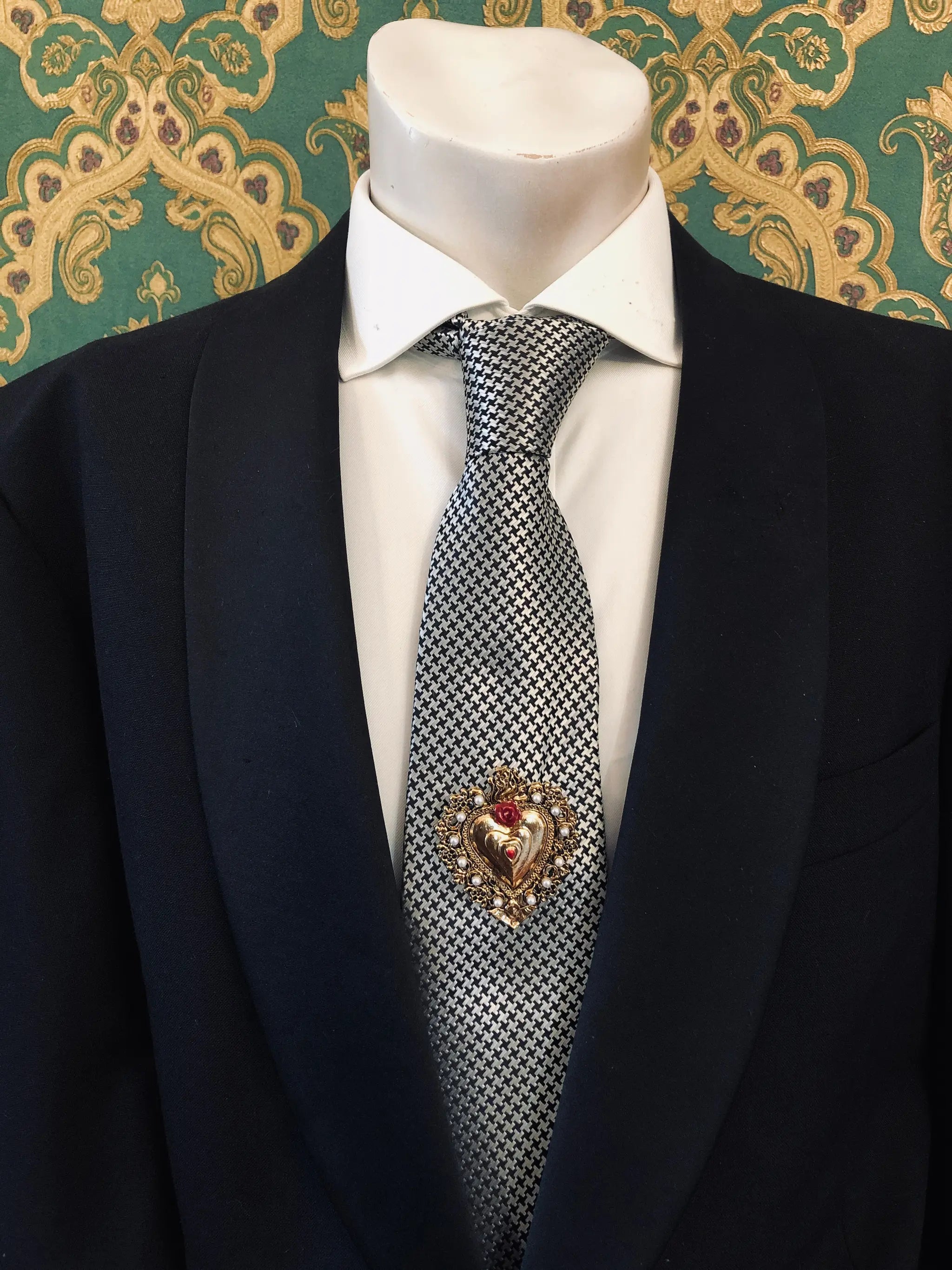 A men's formal outfit displayed on a white mannequin torso, featuring a black suit jacket, a white dress shirt and a patterned grey and white tie. Pinned on the lapel is the Broochella Men's Sacro Cuore collection Benedetta gold heart-shaped brooch with a red flower adornment. The background is a green damask patterned wallpaper.