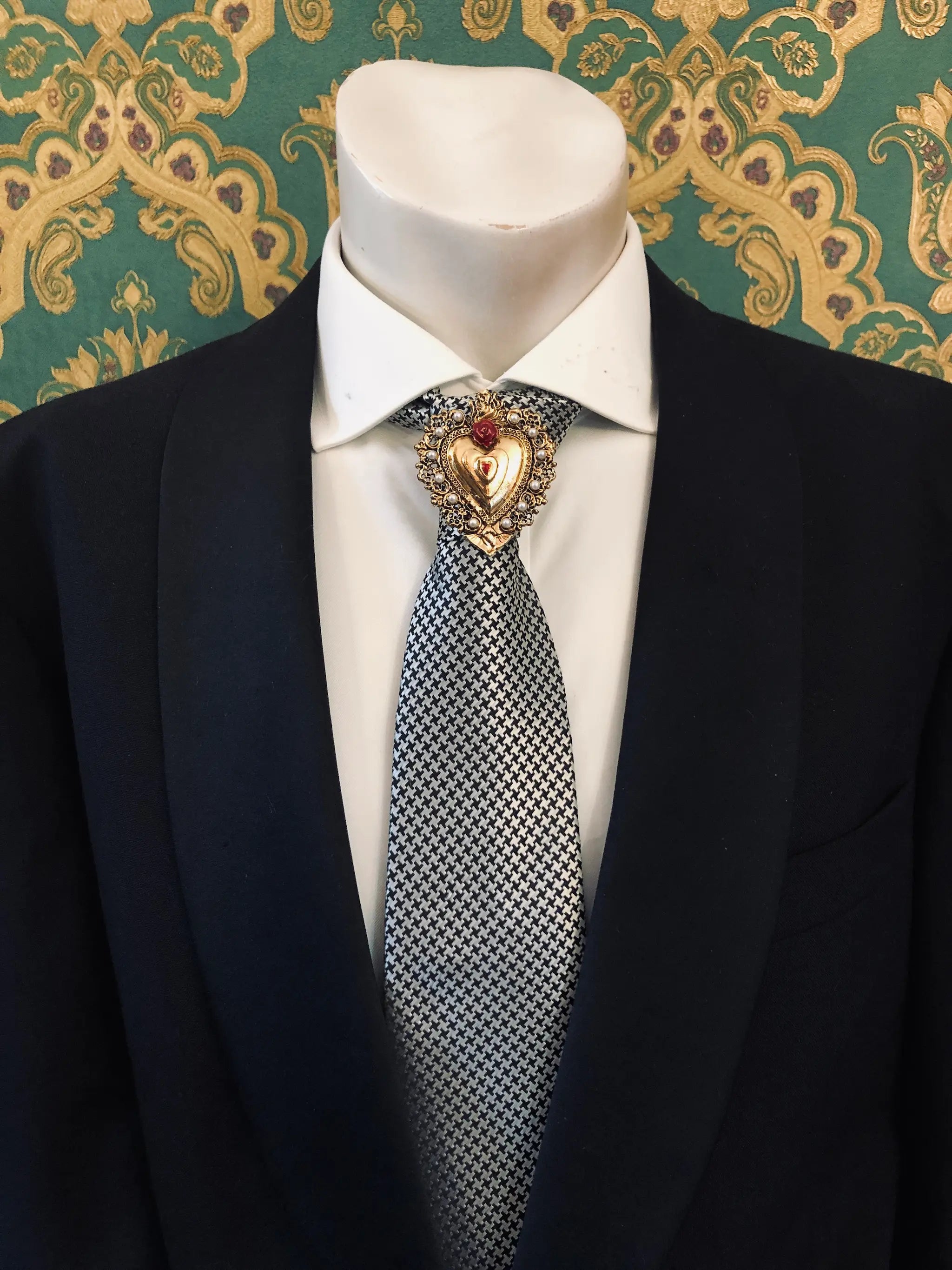 A men's formal outfit displayed on a white mannequin torso, featuring a black suit jacket, a white dress shirt and a patterned grey and white tie. Pinned on the lapel is the Broochella Men's Sacro Cuore collection Benedetta gold heart-shaped brooch with a red flower adornment. The background is a green damask patterned wallpaper.