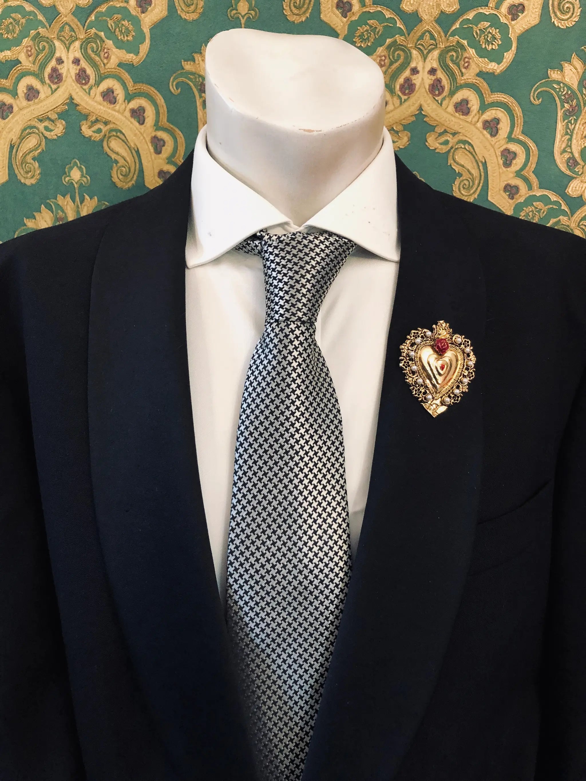 A men's formal outfit displayed on a white mannequin torso, featuring a black suit jacket, a white dress shirt and a patterned grey and white tie. Pinned on the lapel is the Broochella Men's Sacro Cuore collection Benedetta gold heart-shaped brooch with a red flower adornment. The background is a green damask patterned wallpaper.