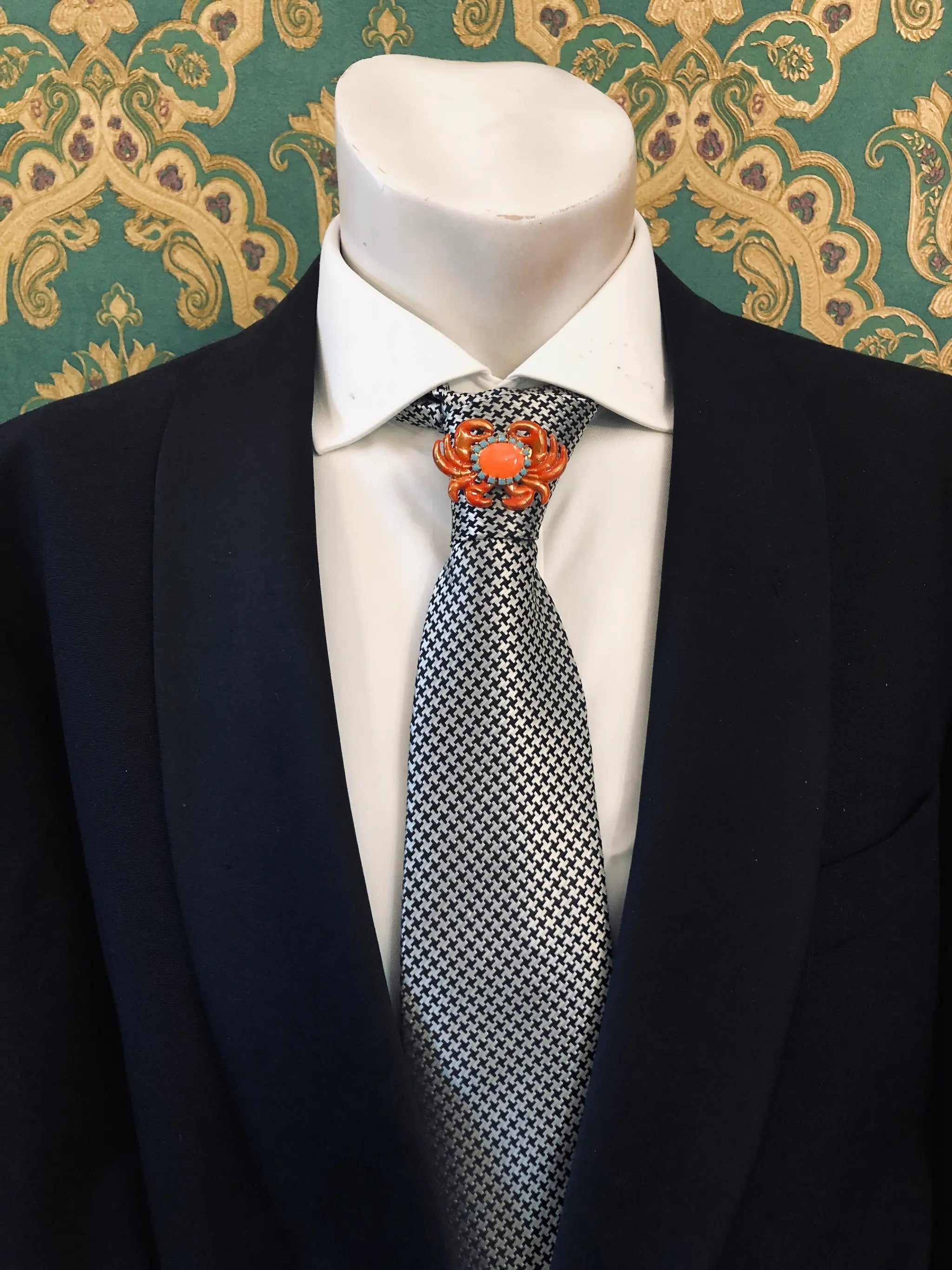 A close-up of a mannequin torso wearing a black suit jacket, white dress shirt, and a black-and-white patterned tie. A Broochella Men's Sacro Cuore  small orange crab-shaped lapel brooch is pinned on the left chest.