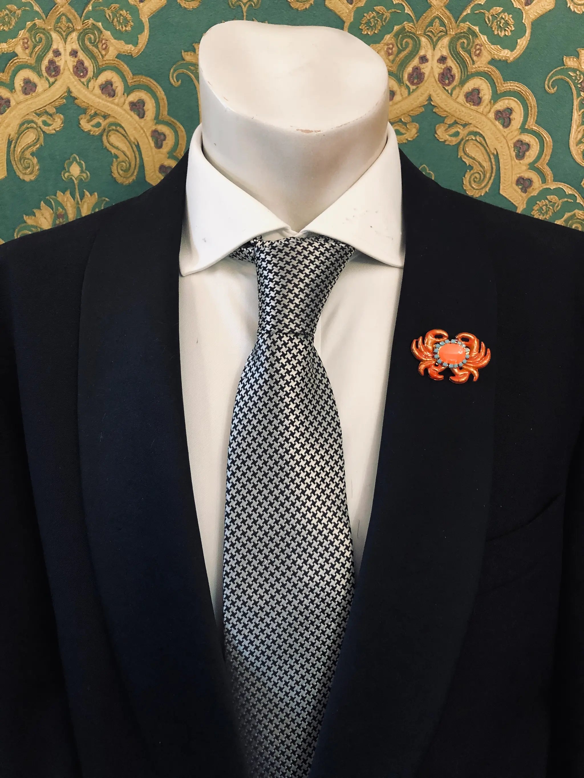 A close-up of a mannequin torso wearing a black suit jacket, white dress shirt, and a black-and-white patterned tie. A Broochella Men's Sacro Cuore  small orange crab-shaped lapel brooch is pinned on the left chest.