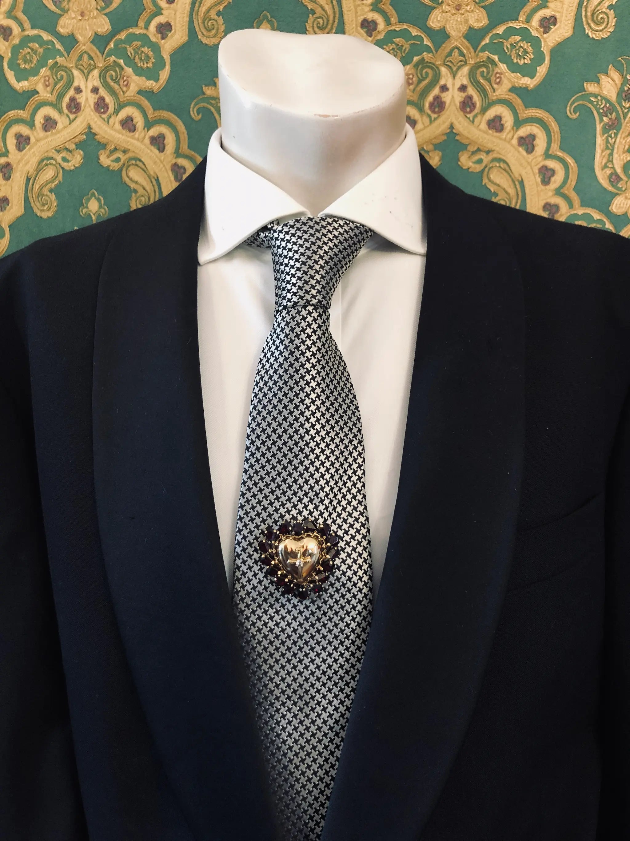A men's necktie with a black and white micro-checkered pattern, styled on a white dress shirt and black suit jacket. A Broochella Men's Sacro Cuore gold heart shaped brooch with crucifix and red stones is pinned on the tie