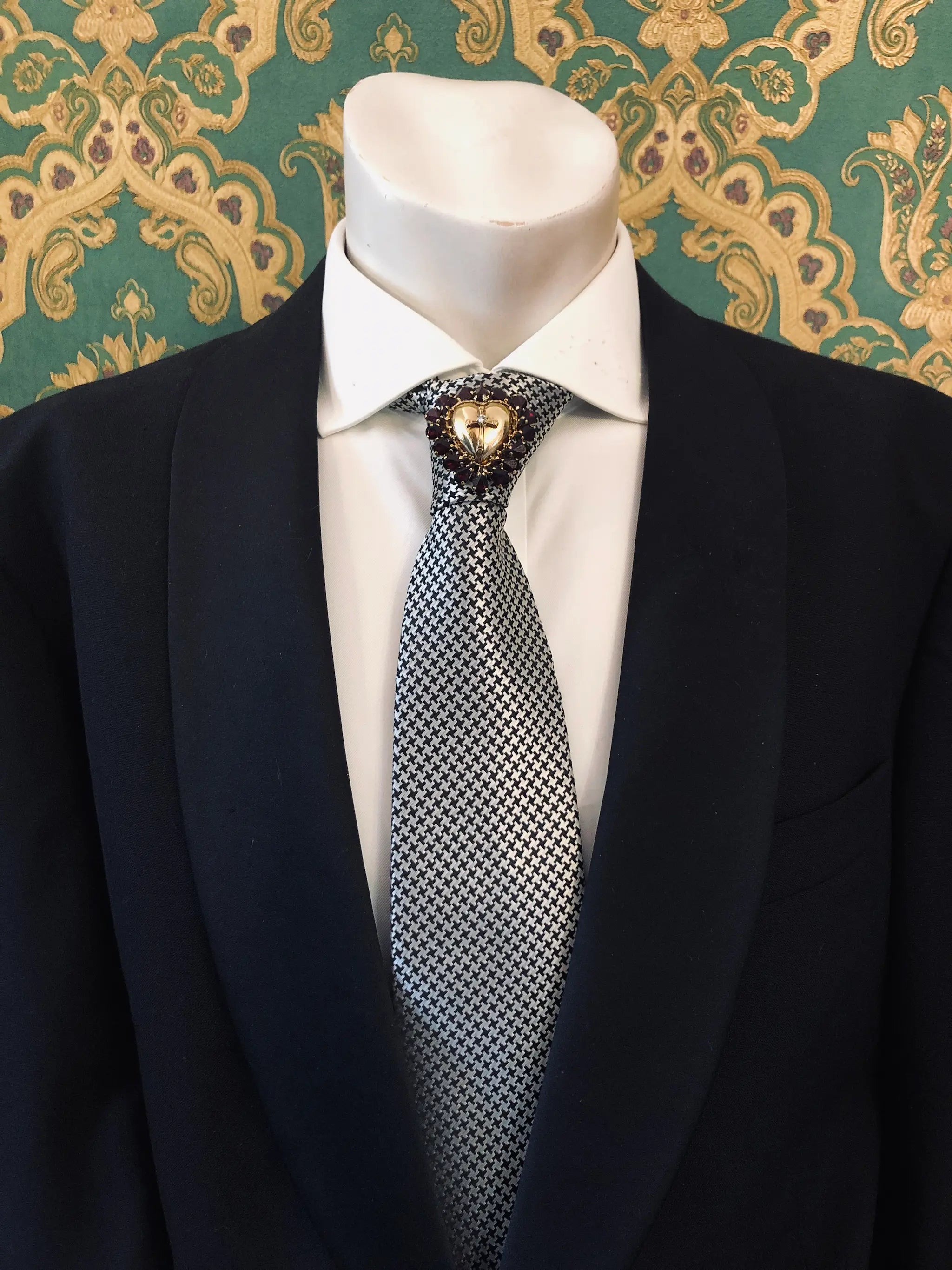 A men's necktie with a black and white micro-checkered pattern, styled on a white dress shirt and black suit jacket. A Broochella Men's Sacro Cuore gold heart shaped brooch with crucifix and red stones is pinned at the collar.