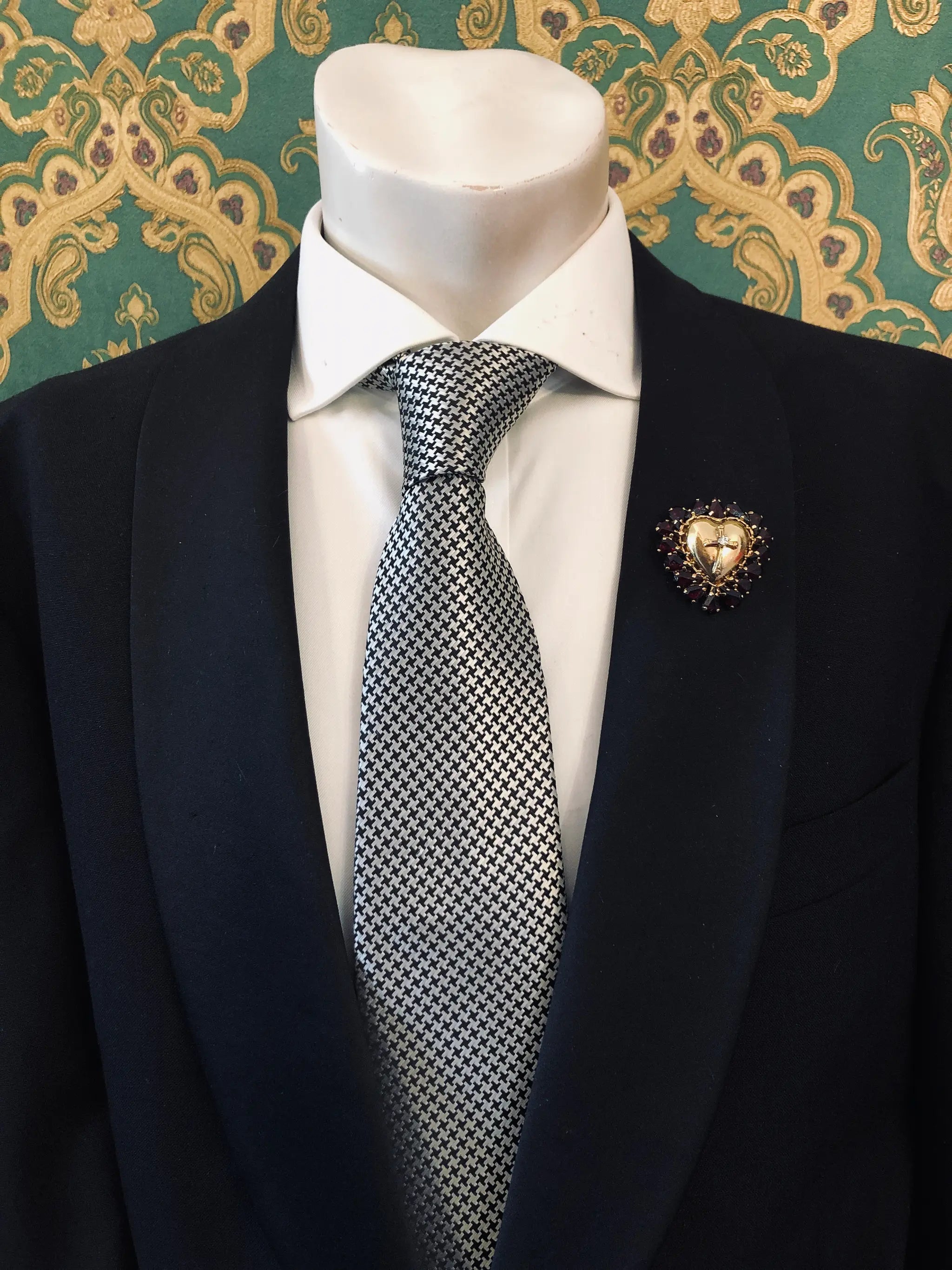 A men's necktie with a black and white micro-checkered pattern, styled on a white dress shirt and black suit jacket. A Broochella Men's Sacro Cuore gold heart shaped brooch with crucifix and red stones is pinned on the lapel