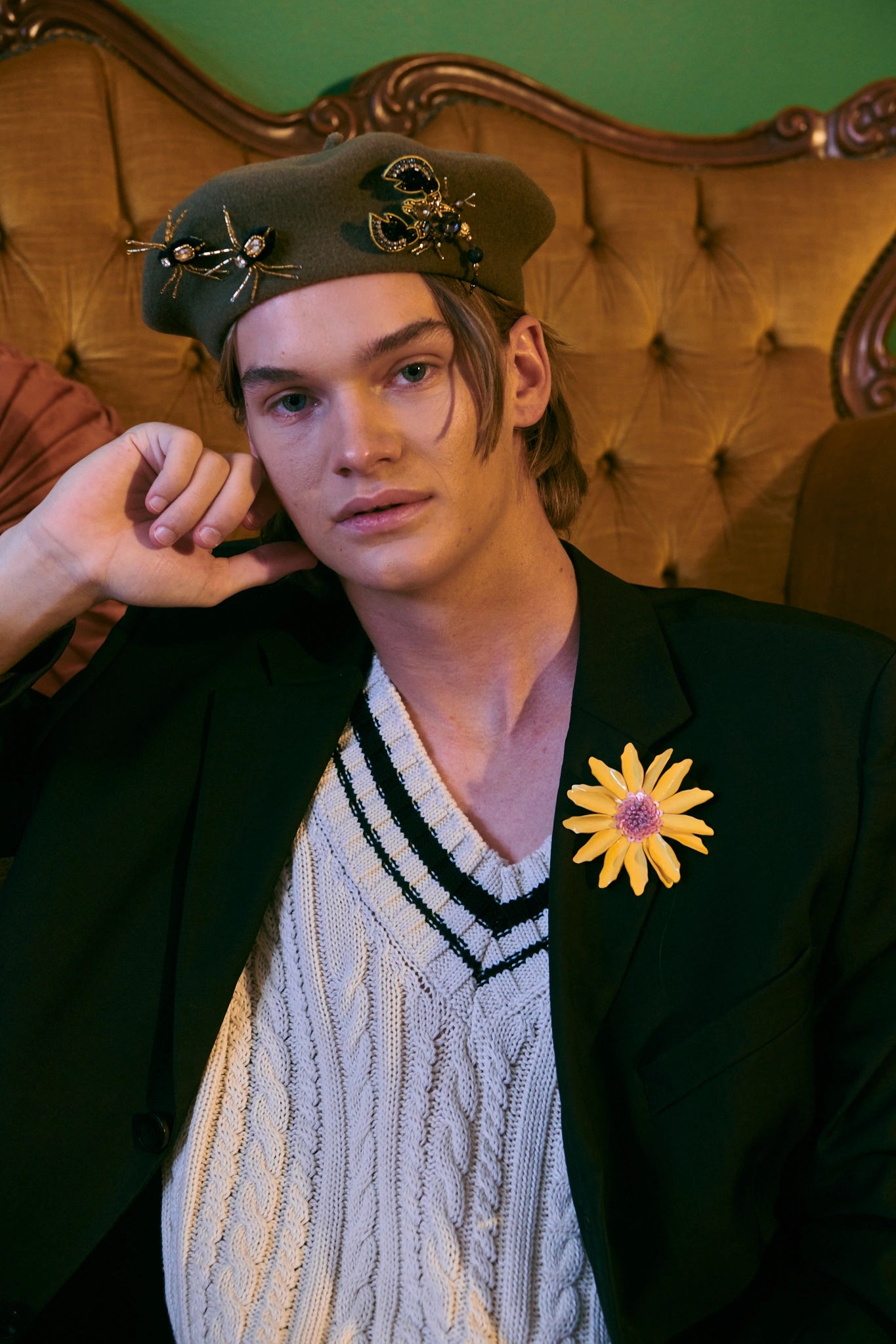 A man wearing a dark olive green beret with Broochella  gold and black scorpion embellishments, a black blazer, and a cream cable-knit sweater vest, seated in front of a tufted brown leather sofa. He has a yellow flower pin on his blazer.